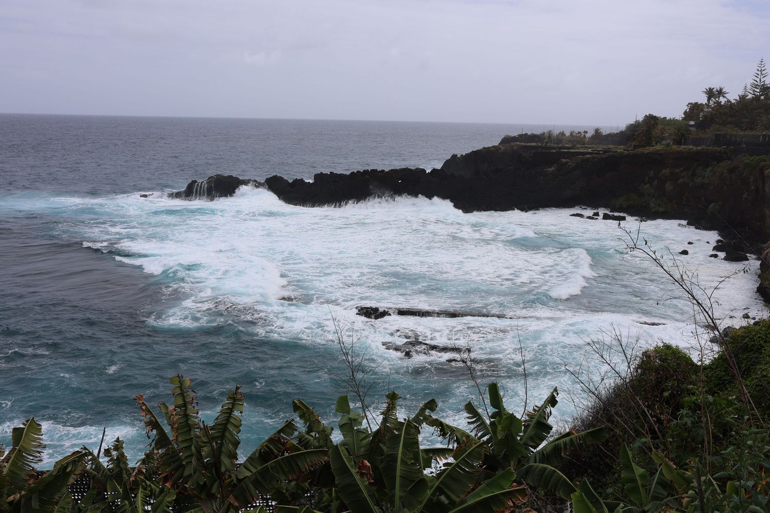 Dramatic ocean waves crashing against volcanic black rocks along a rugged tropical coastline under overcast skies.