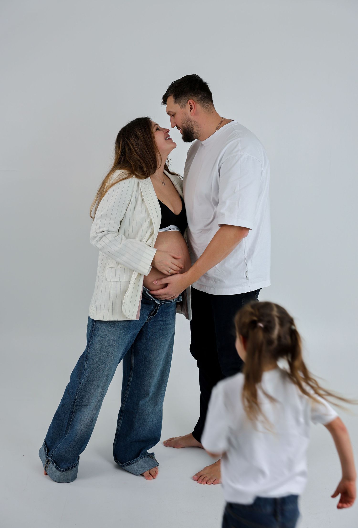 Pregnant couple sharing a tender moment while a young girl plays in the foreground during a maternity photoshoot.