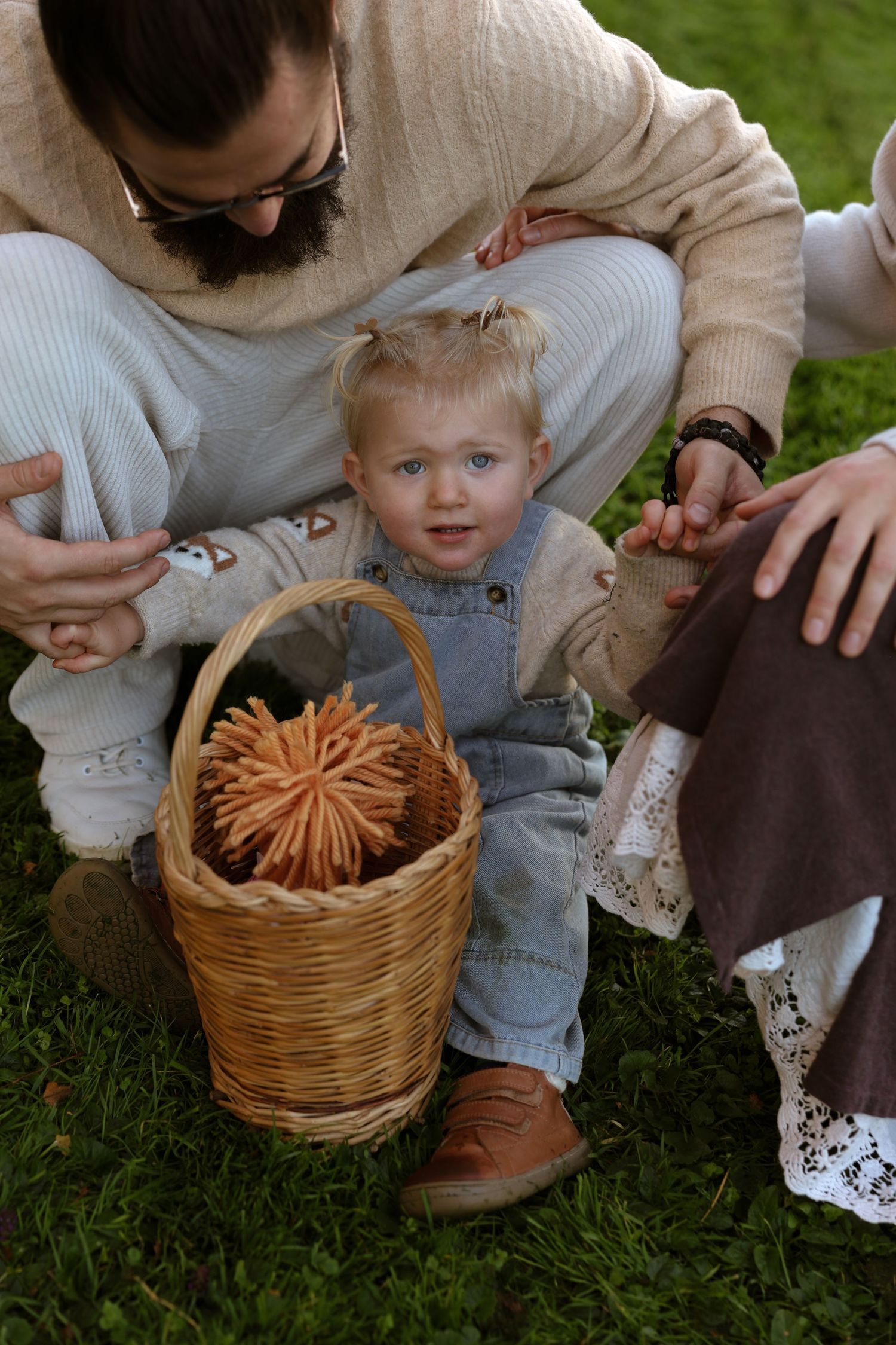 Toddler in denim overalls stands beside a wicker basket of cookies on grass, supported by a bearded man in a cream sweater.