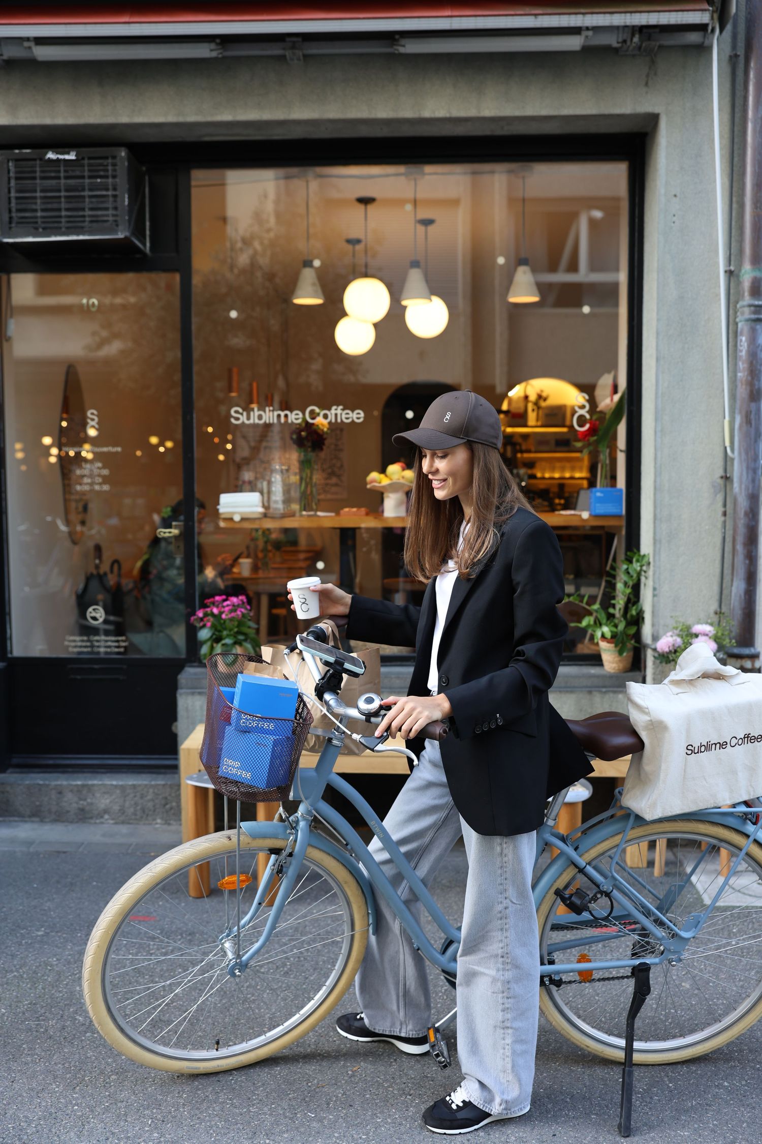 Woman in black blazer and cap stands with vintage blue bike outside Sublime Coffee shop.