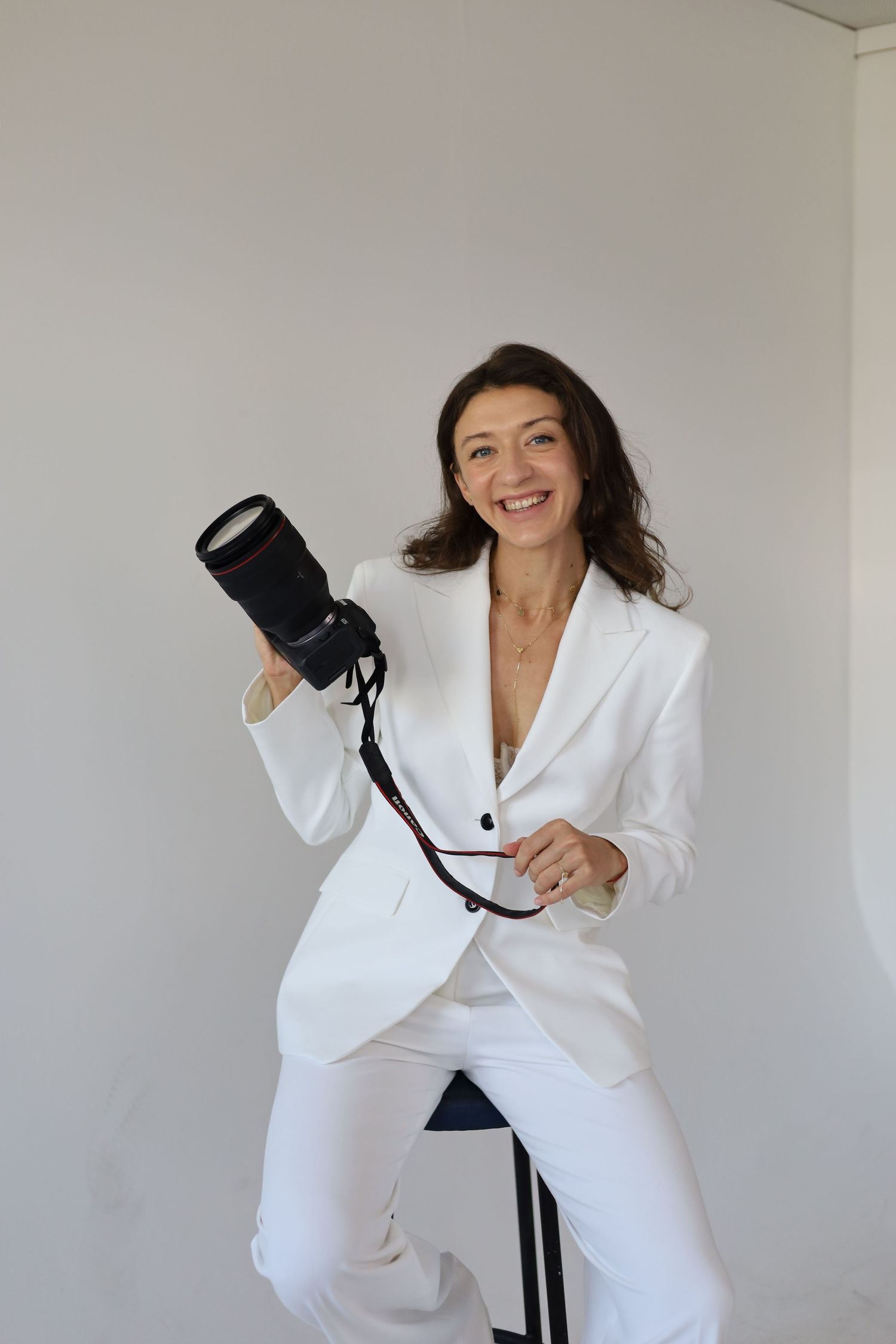 Smiling woman in white suit holding a professional DSLR camera with strap, seated against a light gray background.