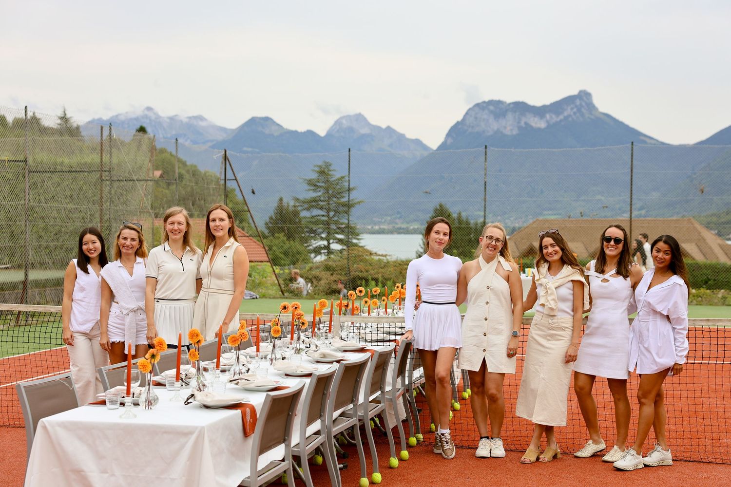 Women in white tennis outfits gather around an elegant outdoor dining table on a court with mountain views.