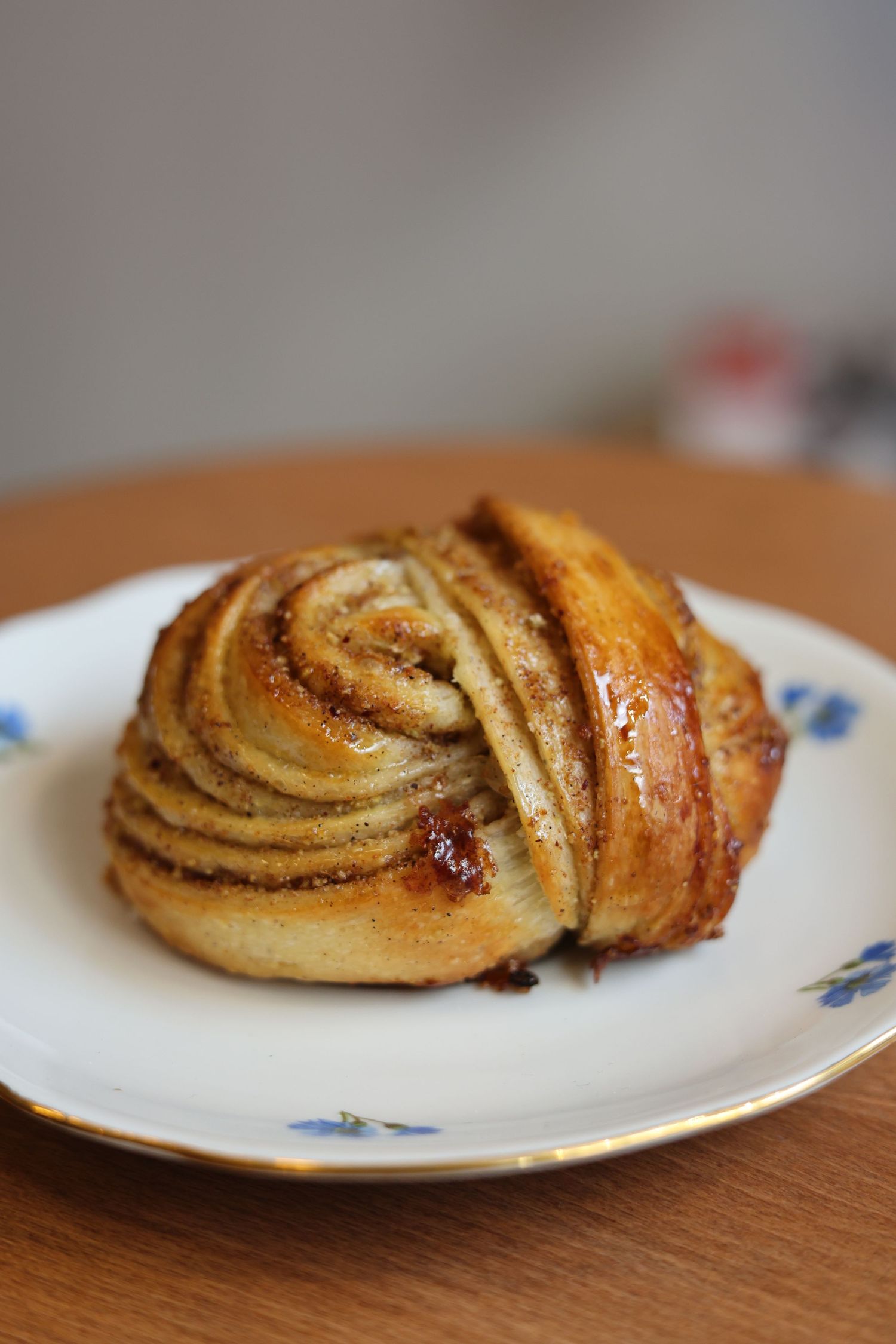 A golden-brown cinnamon swirl pastry on a white plate with blue floral trim, placed on a wooden table.