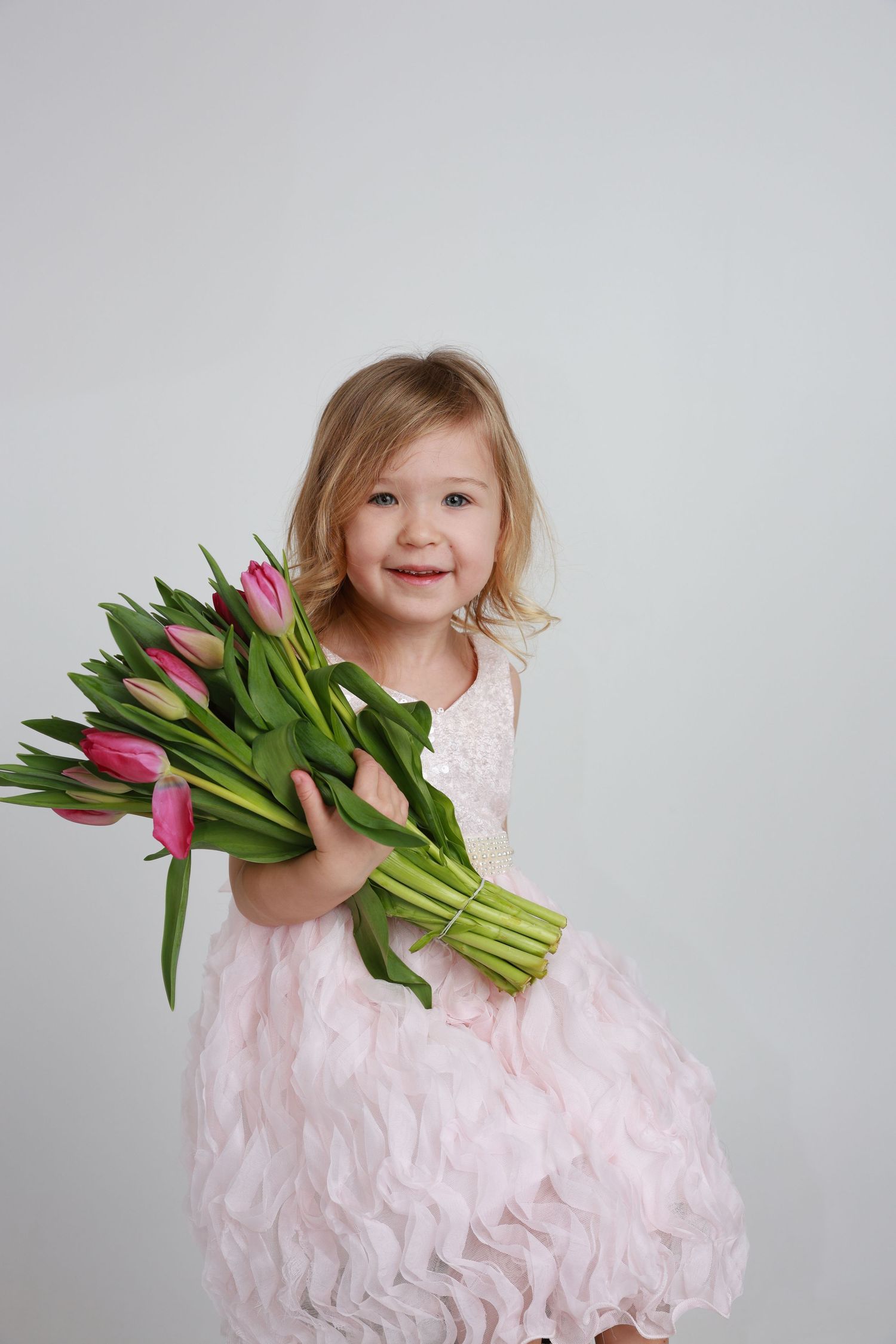 Smiling little girl in pink dress holding bouquet of pink tulips on white background.
