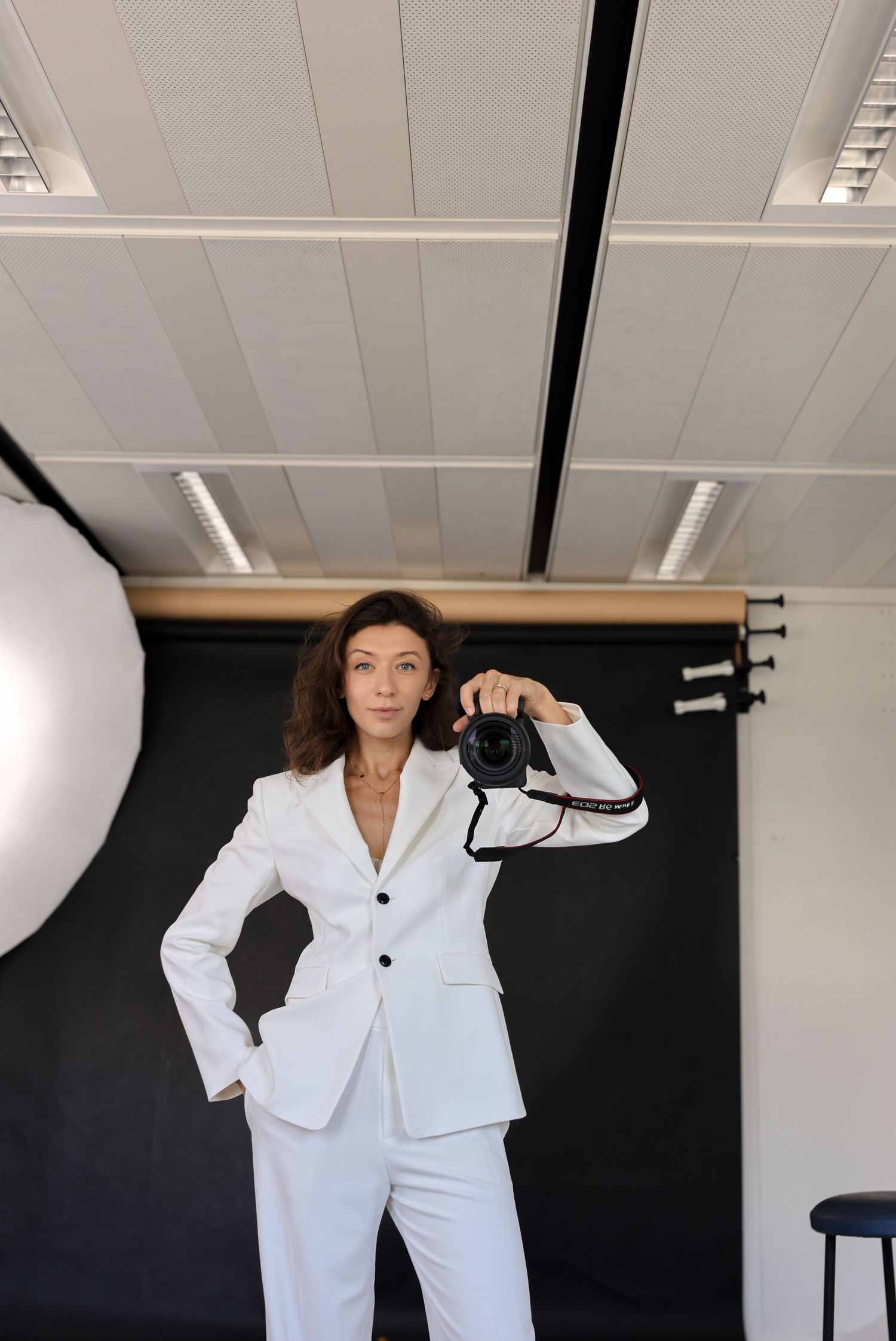 Woman in white suit holding a professional camera in a photography studio with black backdrop.