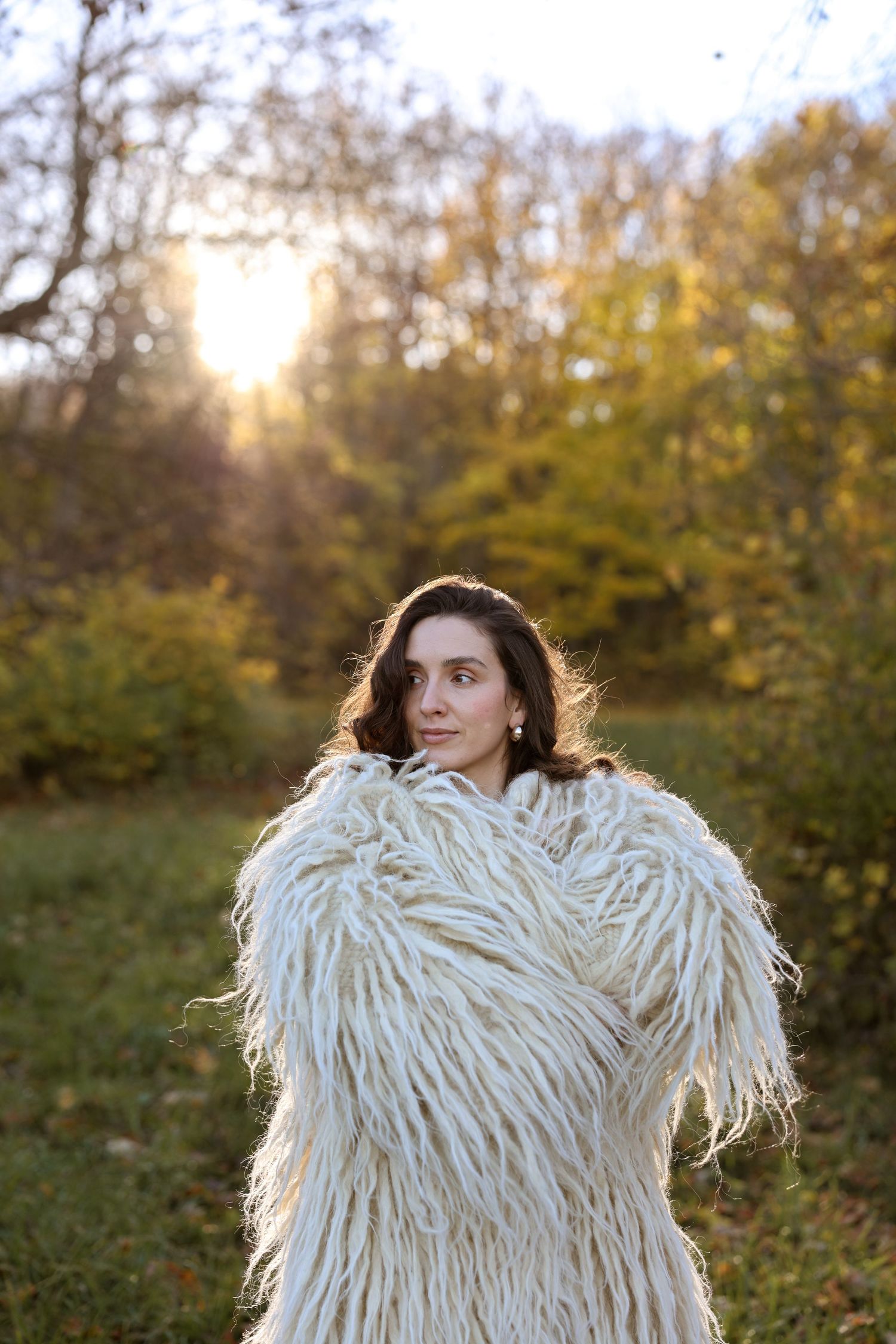 Woman in white fluffy textured coat standing in autumn park with golden sunlight filtering through colorful trees.