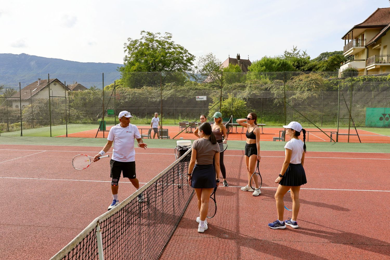 Group tennis lesson on outdoor red clay court with coach and students near the net on a sunny day.