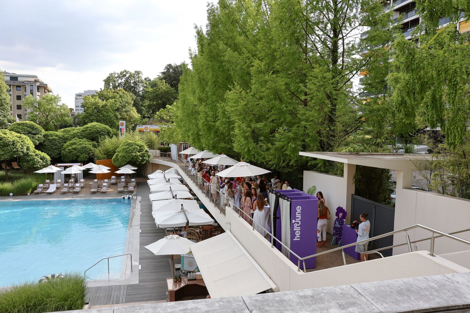 Outdoor poolside event with white umbrellas, lush green trees, and guests gathered near a purple branded display booth.