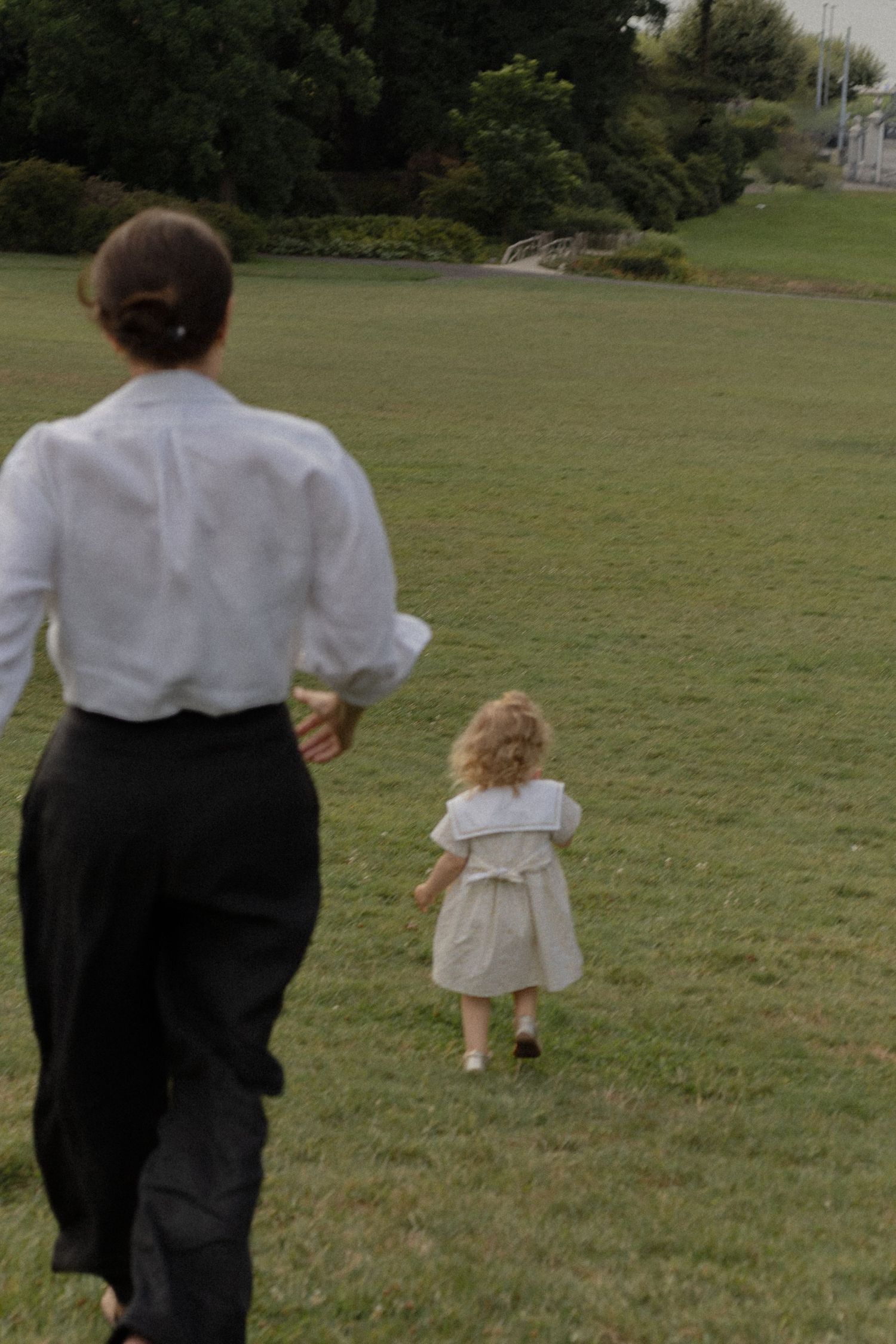 Woman in white blouse and black skirt walks with a young girl in a white dress across a green grassy park.