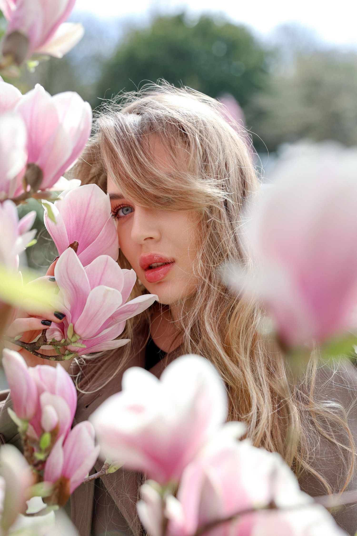 Young blonde woman with wavy hair poses among blooming pink magnolia flowers in a soft spring outdoor setting.