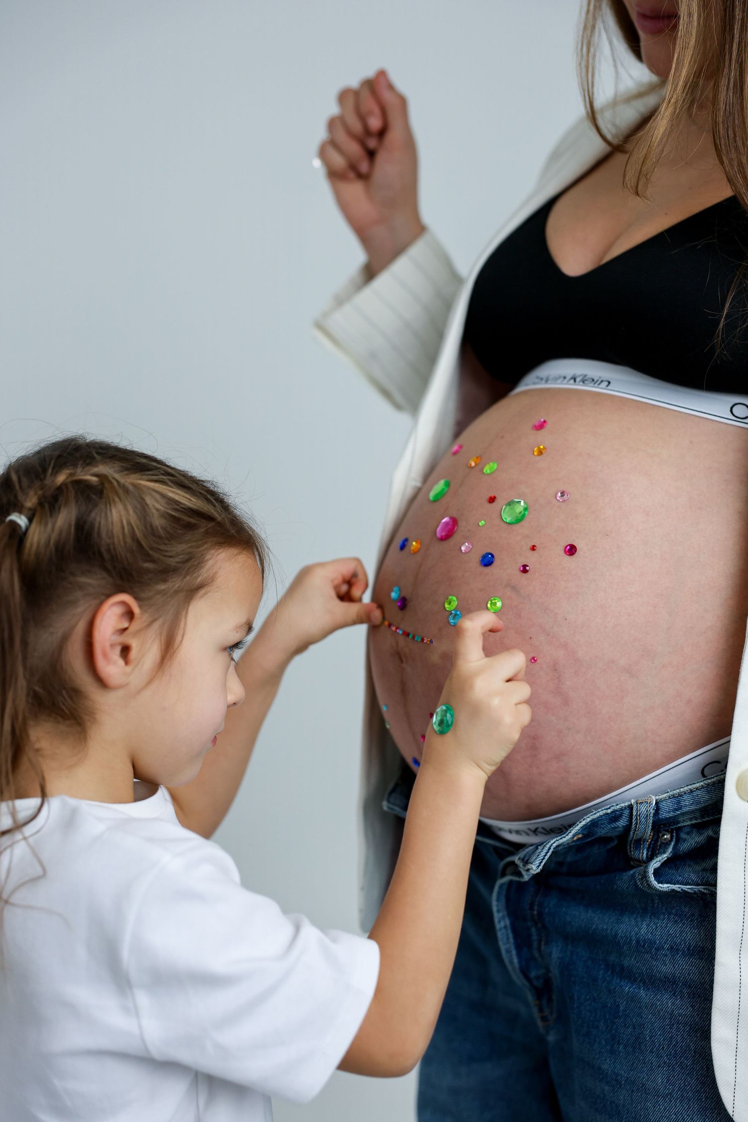 Young girl decorating pregnant woman's belly with colorful gems in a playful maternity photo shoot.