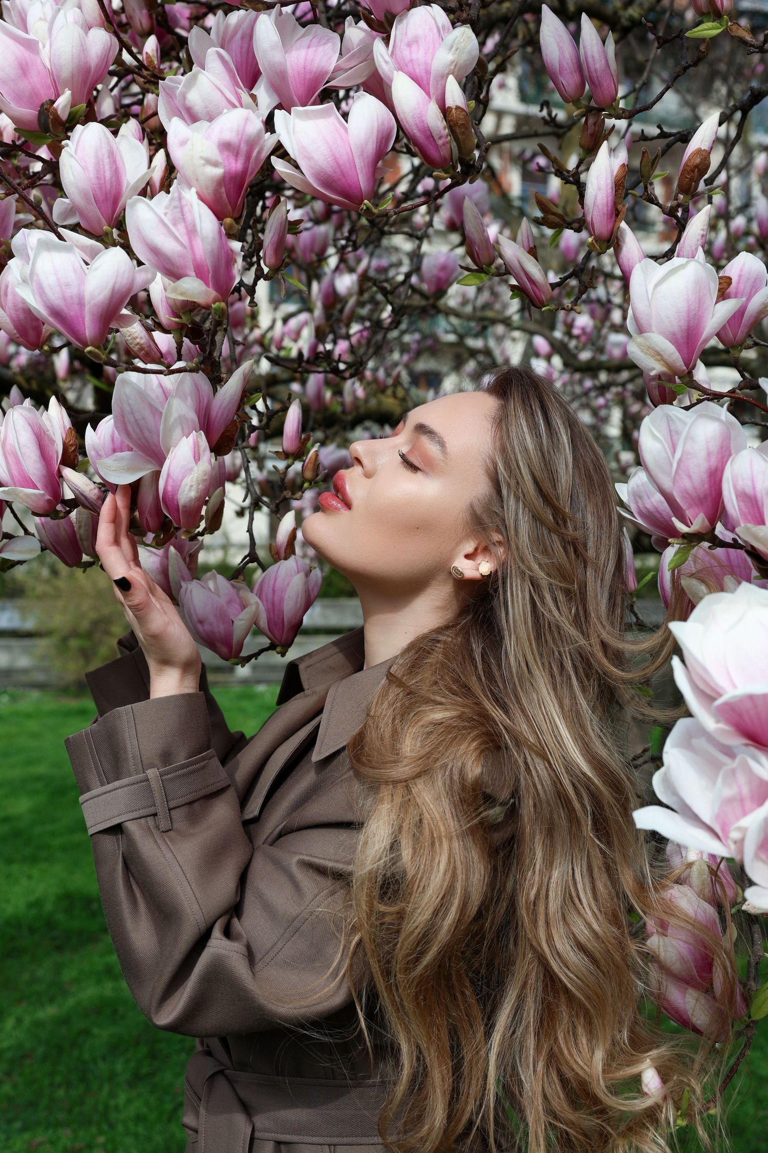 Young woman with long hair in brown leather jacket posing near blooming pink magnolia tree in spring garden.