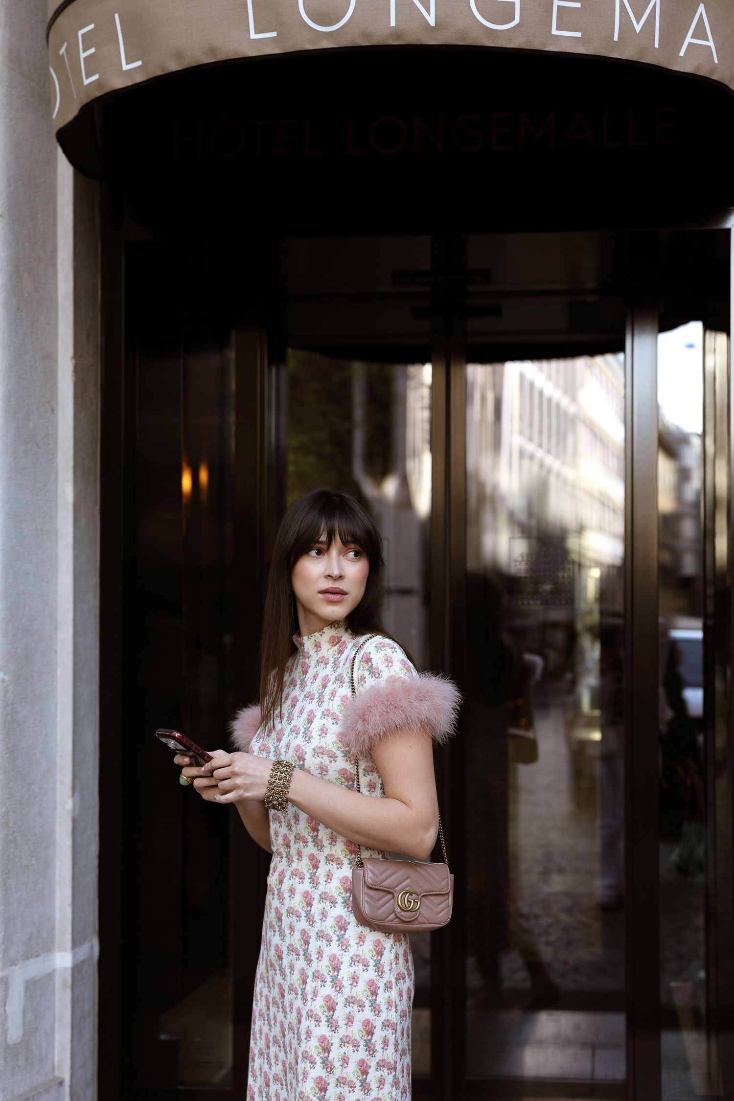 Woman in floral dress with pink feather trim holds phone outside Hotel Longema, carrying a Gucci bag.