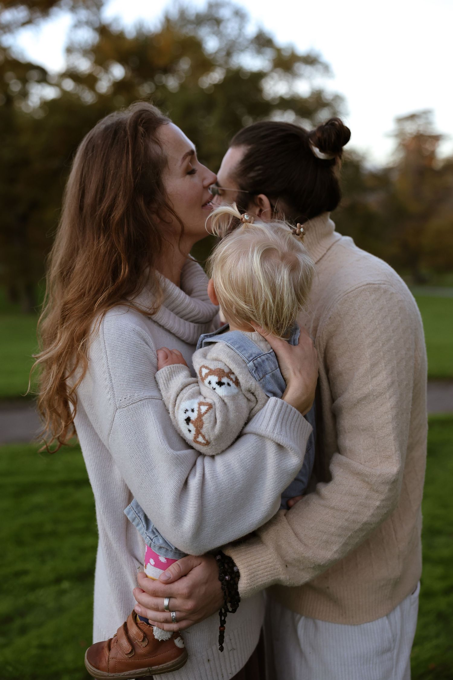 Two women sharing a kiss while holding a young child in a park during autumn.
