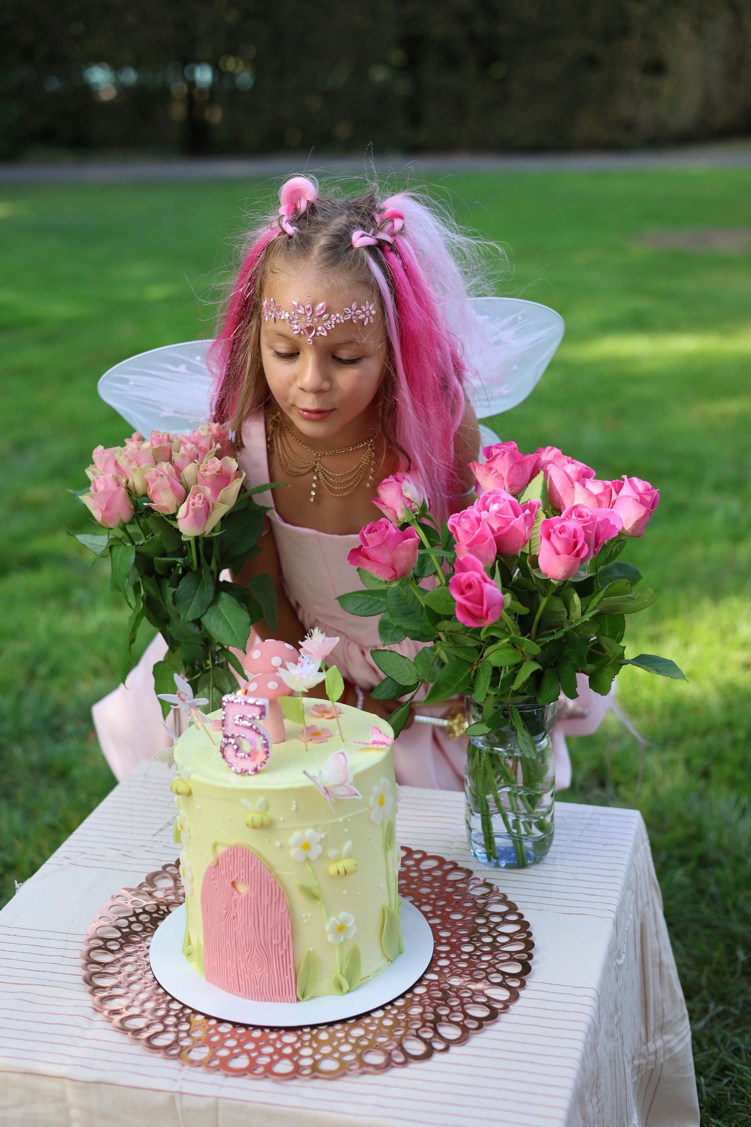 Girl in fairy costume with pink wings celebrates 5th birthday with floral cake and roses outdoors.
