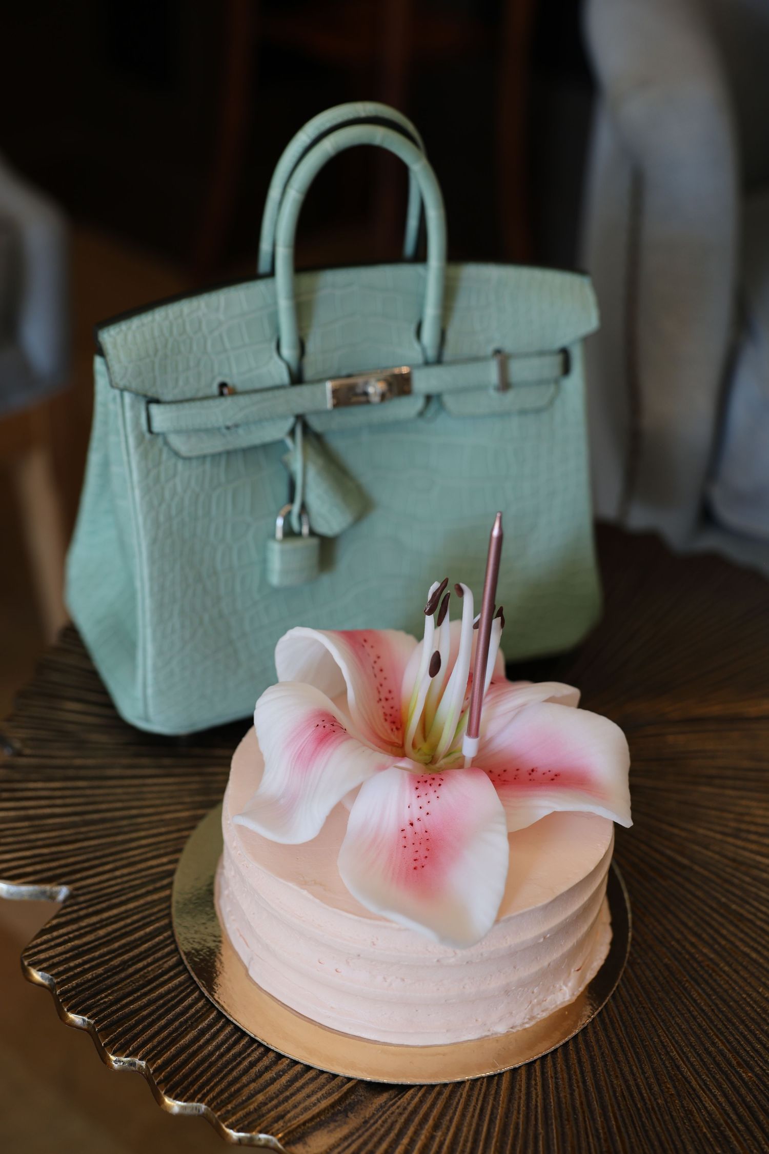 Pink birthday cake with lily flower and candle displayed in front of a mint green Hermes Birkin bag on gold tray.