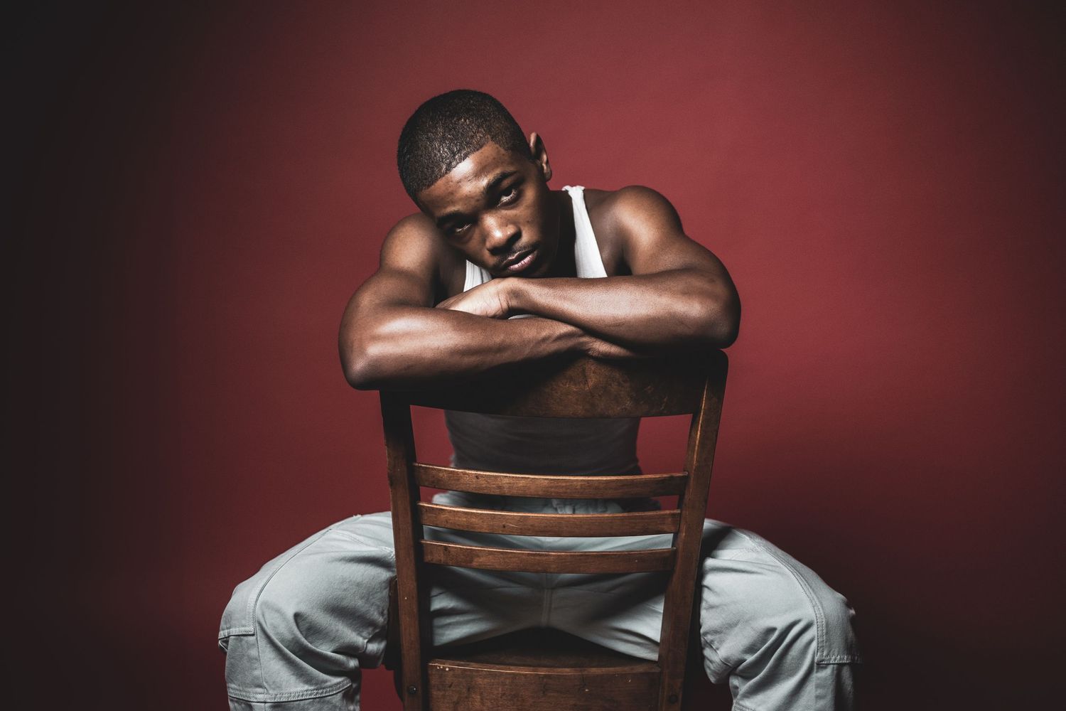Dark silhouette of a person sitting on a wooden chair in a dramatic red-lit setting wearing a white tank top.