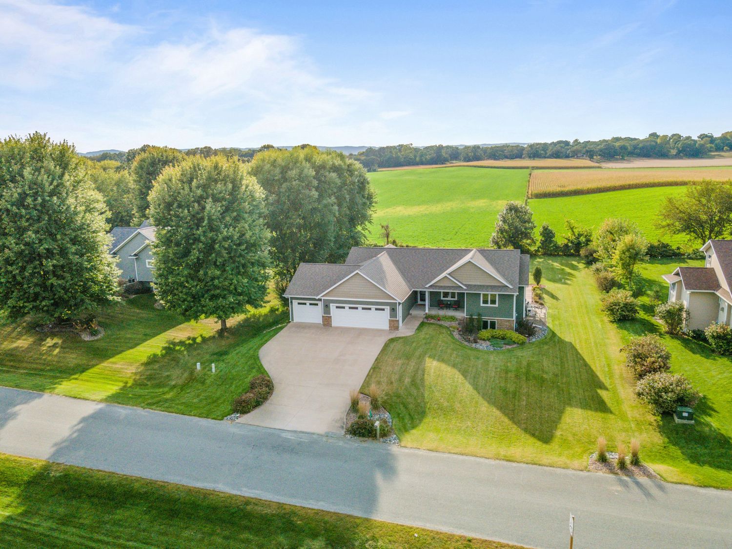 Aerial view of a modern ranch-style home with beige siding and attached garage surrounded by green lawns and rural landscape.