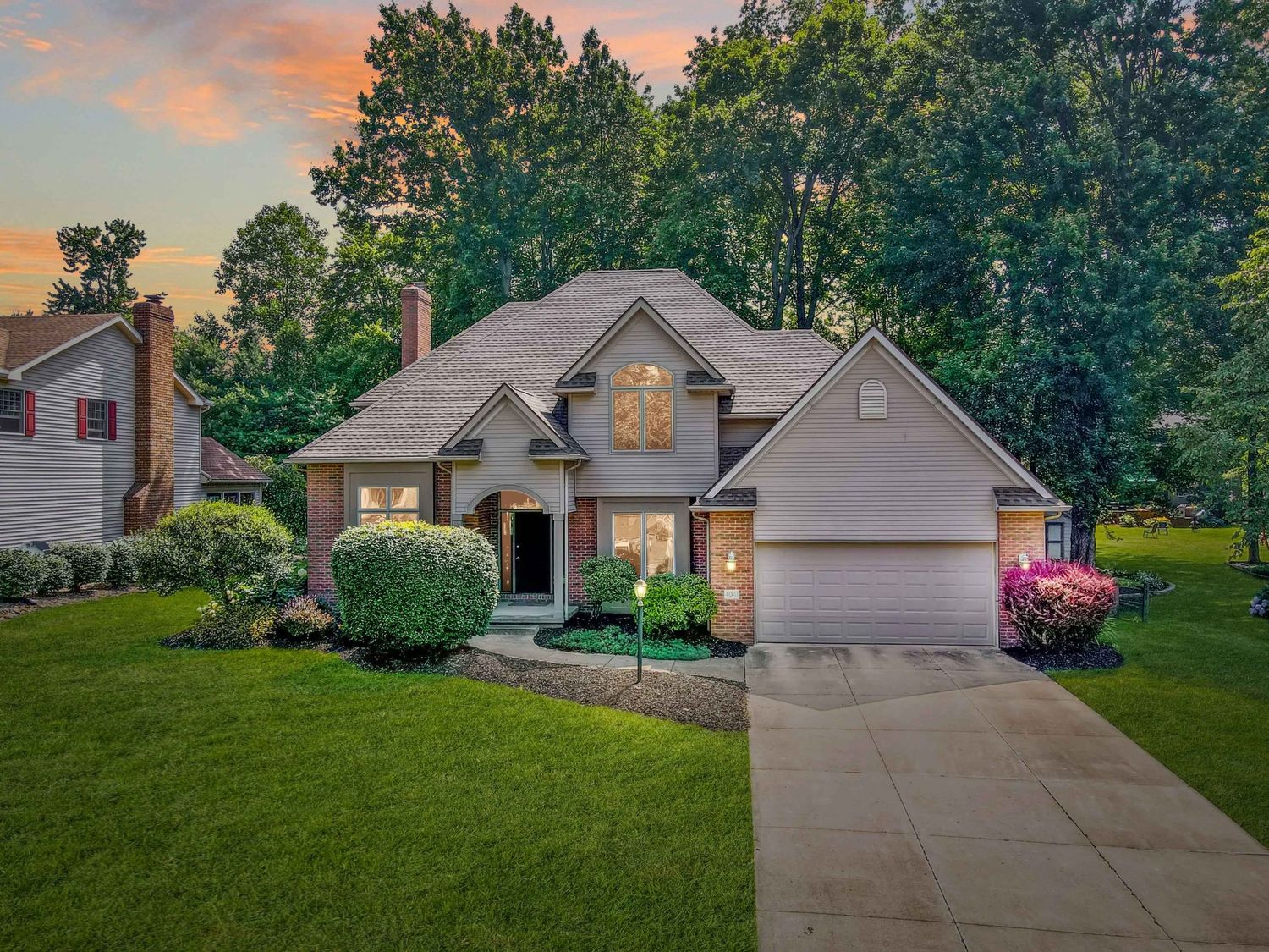 A series of suburban homes with attached garages and driveways surrounded by green lawns and trees at sunset.