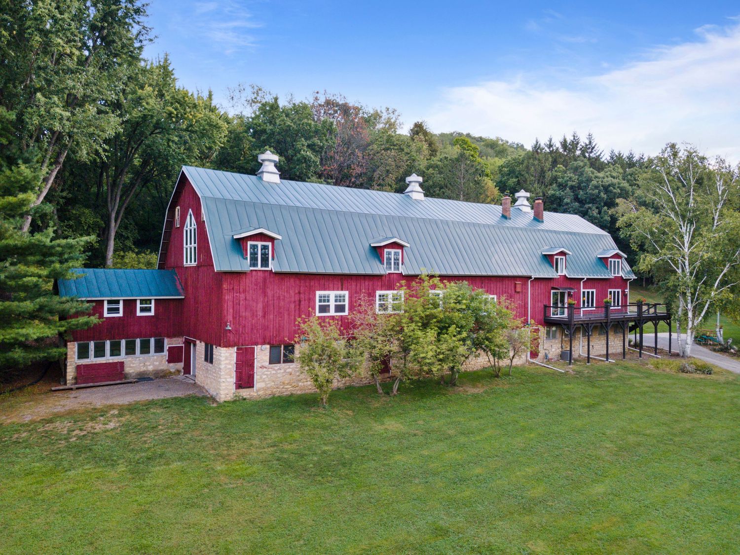 Large red barn-style building with grey metal roof and white trim set on grassy lawn surrounded by trees.