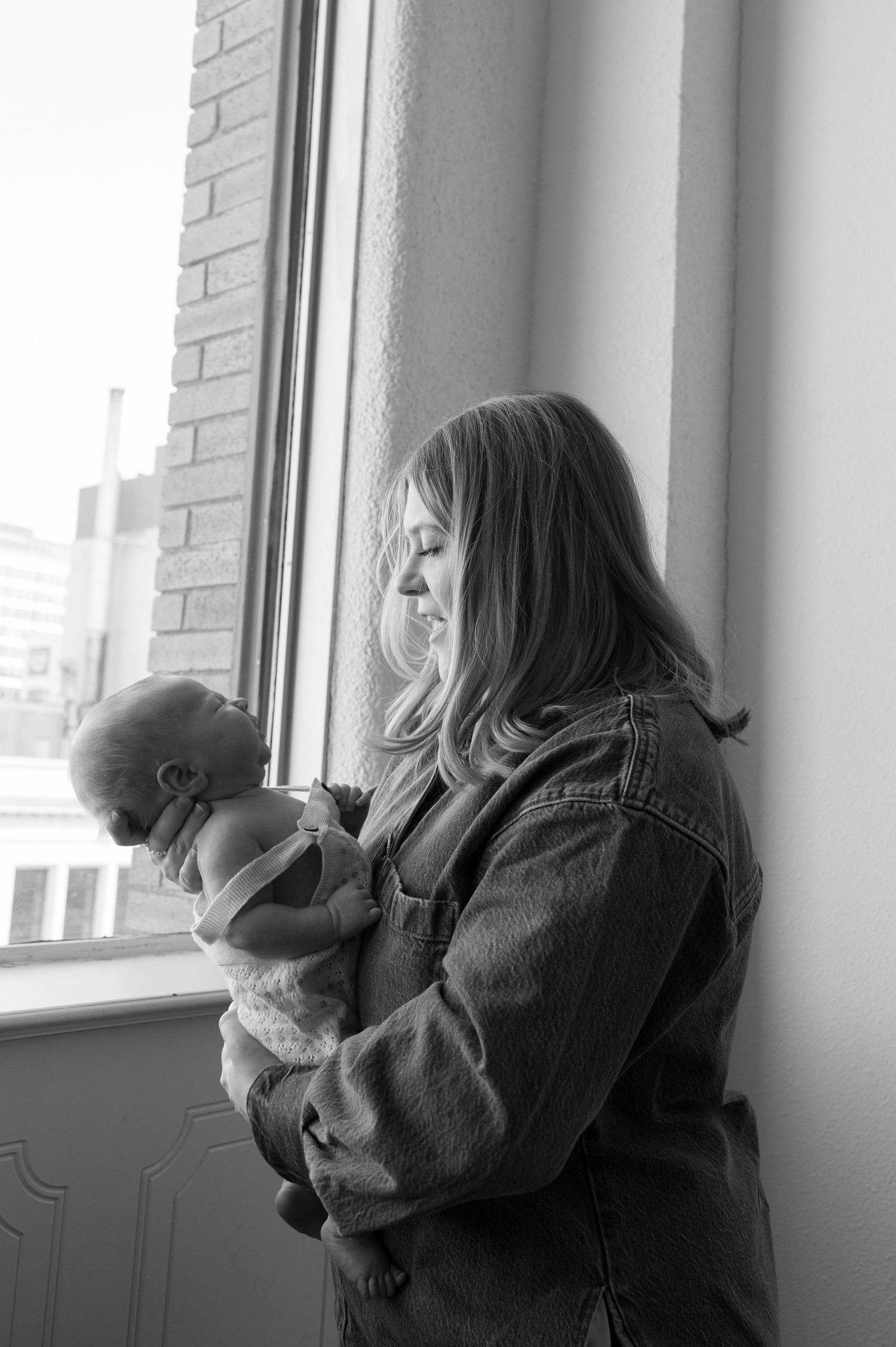 A tender black and white portrait of a parent holding a baby while standing by a window in a denim jacket.