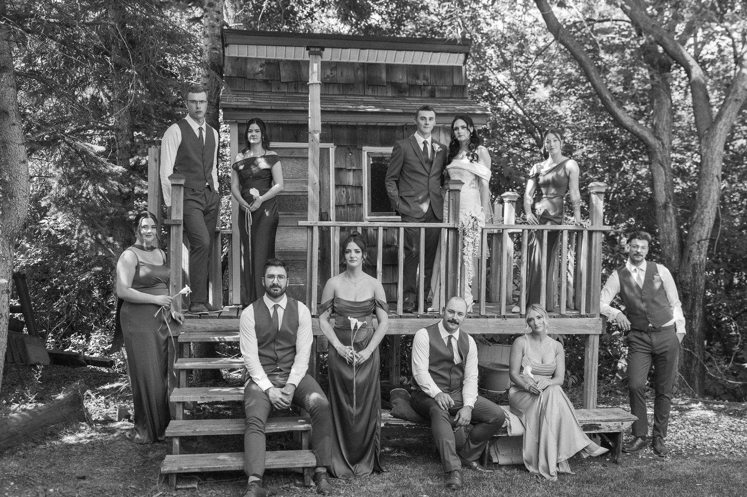 Black and white group portrait of a wedding party in vintage attire posing on the steps and porch of a rustic wooden cabin among trees.