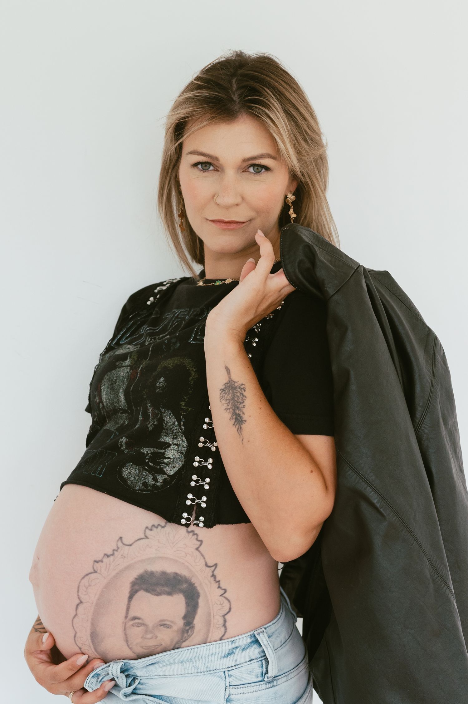 A pregnant person in a black top and denim jeans displays their belly with a portrait tattoo.