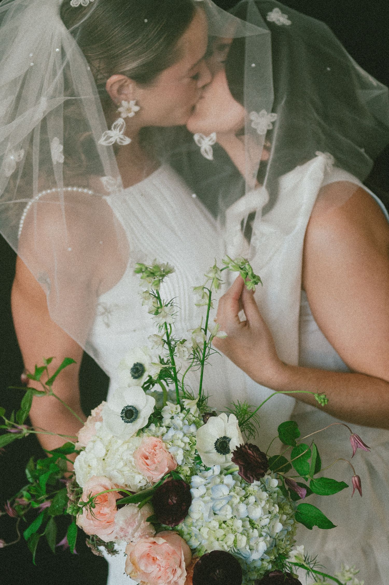 Two brides share a romantic kiss beneath a veil with a pastel floral bouquet.
