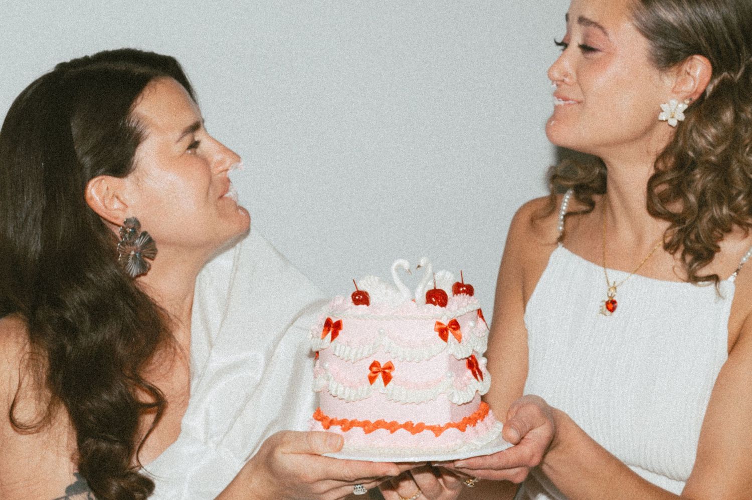 Two people in white dresses hold a white birthday cake decorated with red bows and orange trim together.