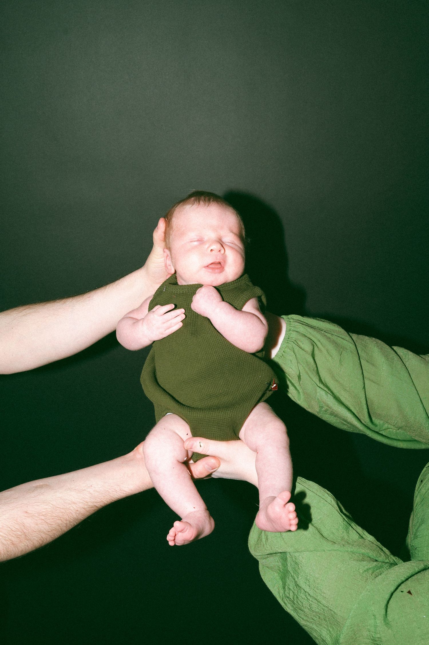A tiny infant in a green onesie being gently supported by adult hands against a dark background.