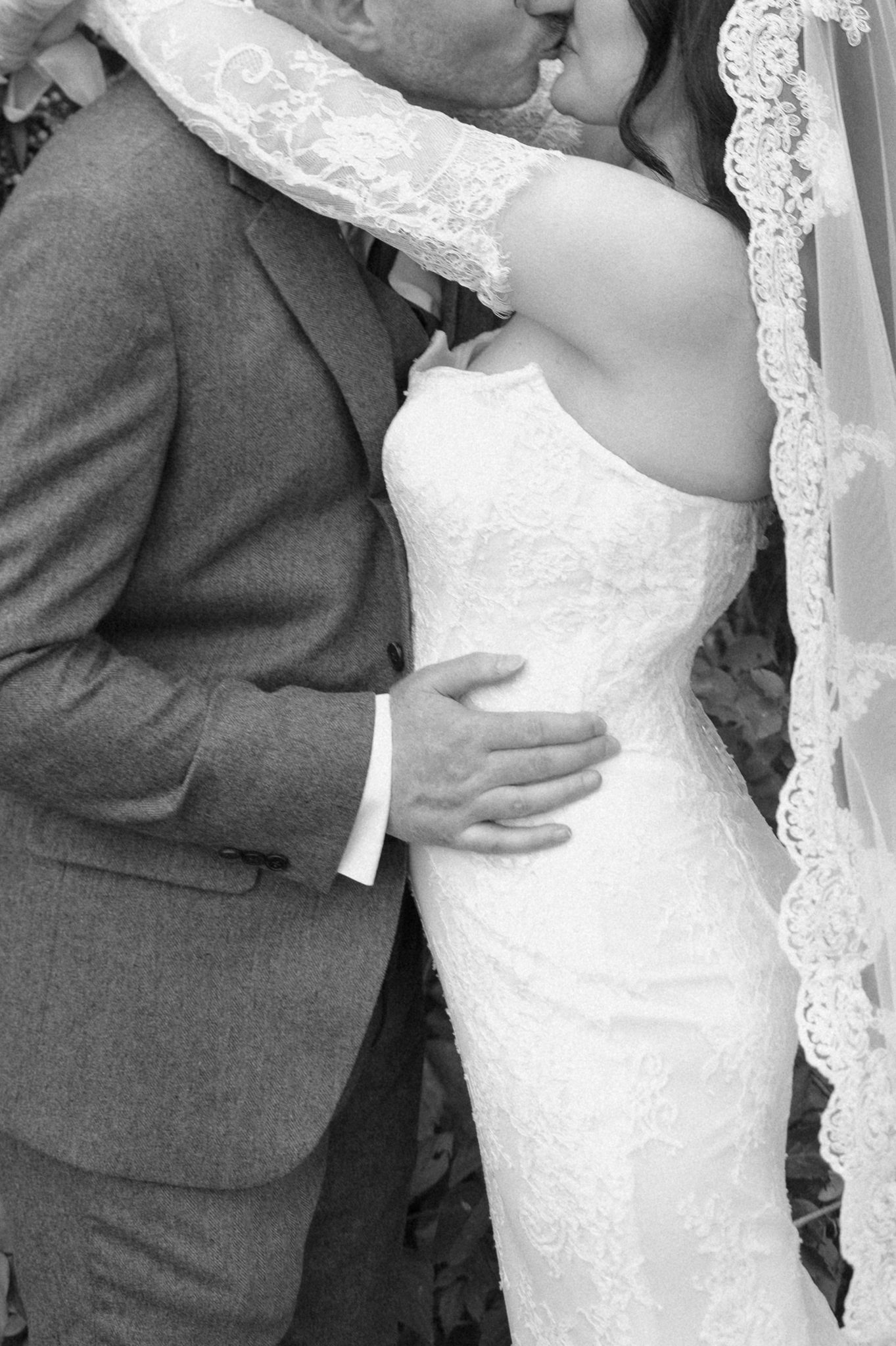 Black and white photo of a bride in a strapless lace gown and veil kissing her groom in a tweed suit, his hand on her waist.