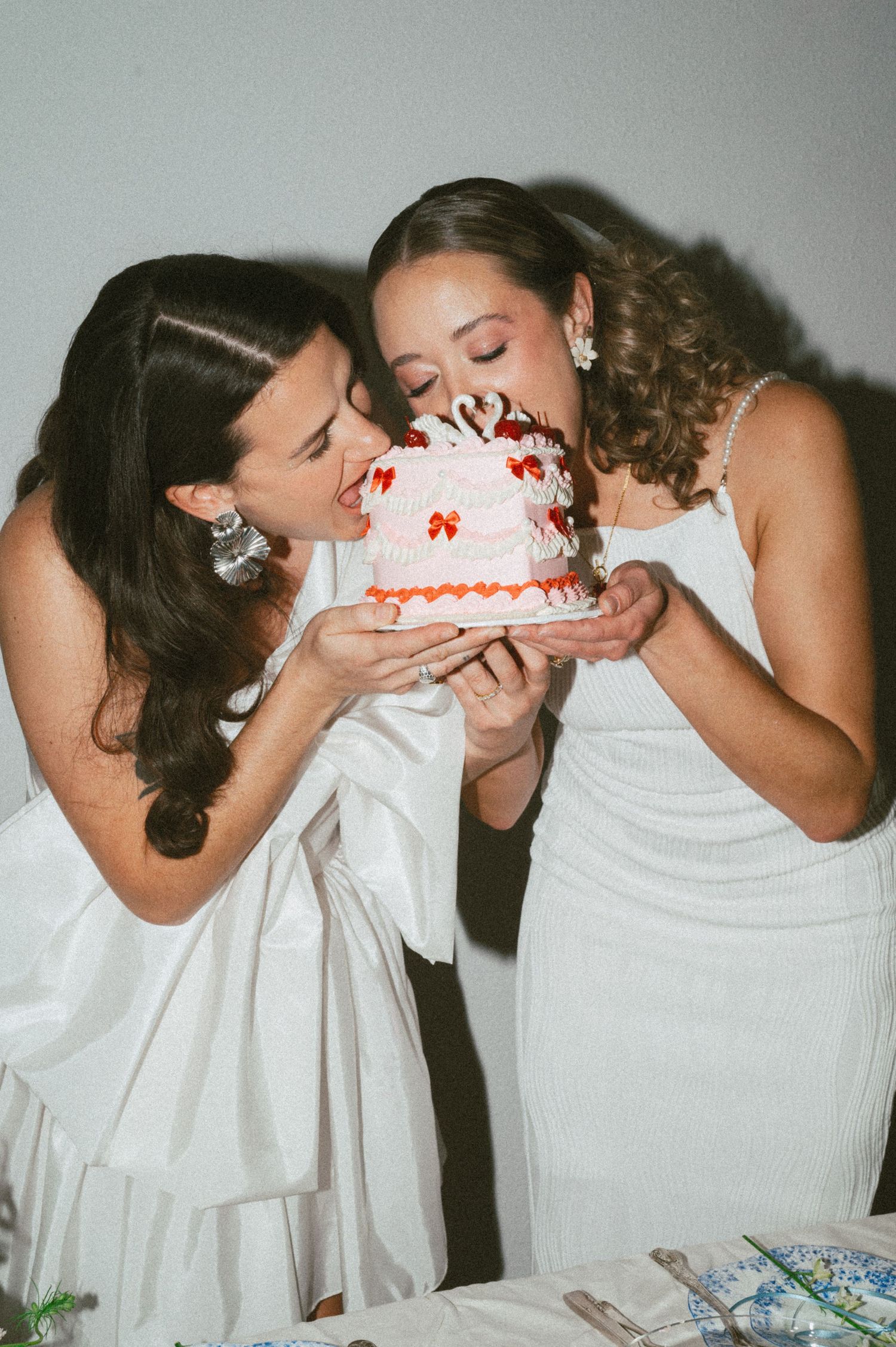 Two women in white dresses playfully taking bites from a decorated pink birthday cake together.