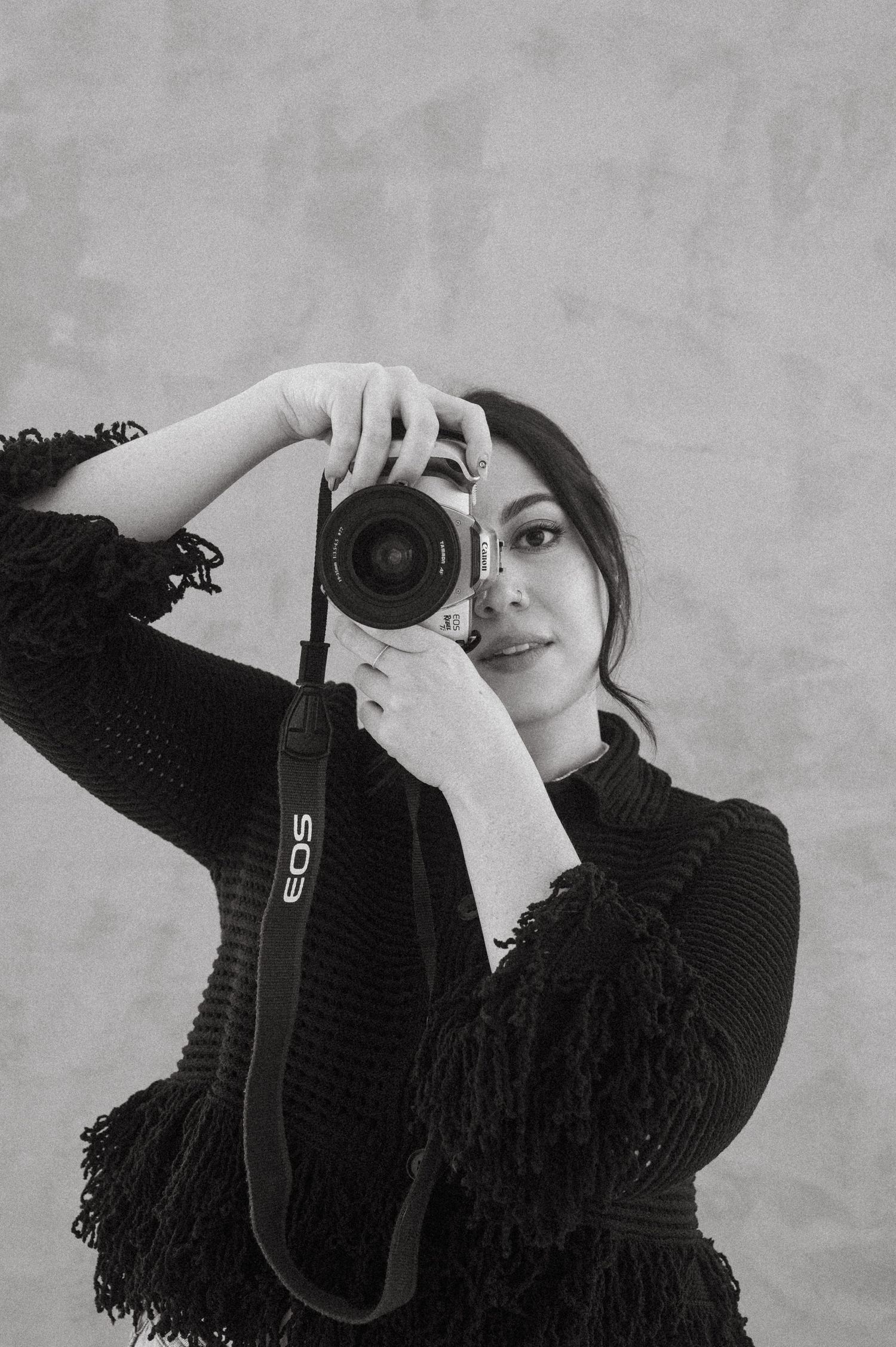 A photographer takes a self-portrait with a DSLR camera while wearing a black sweater against a light gray background.