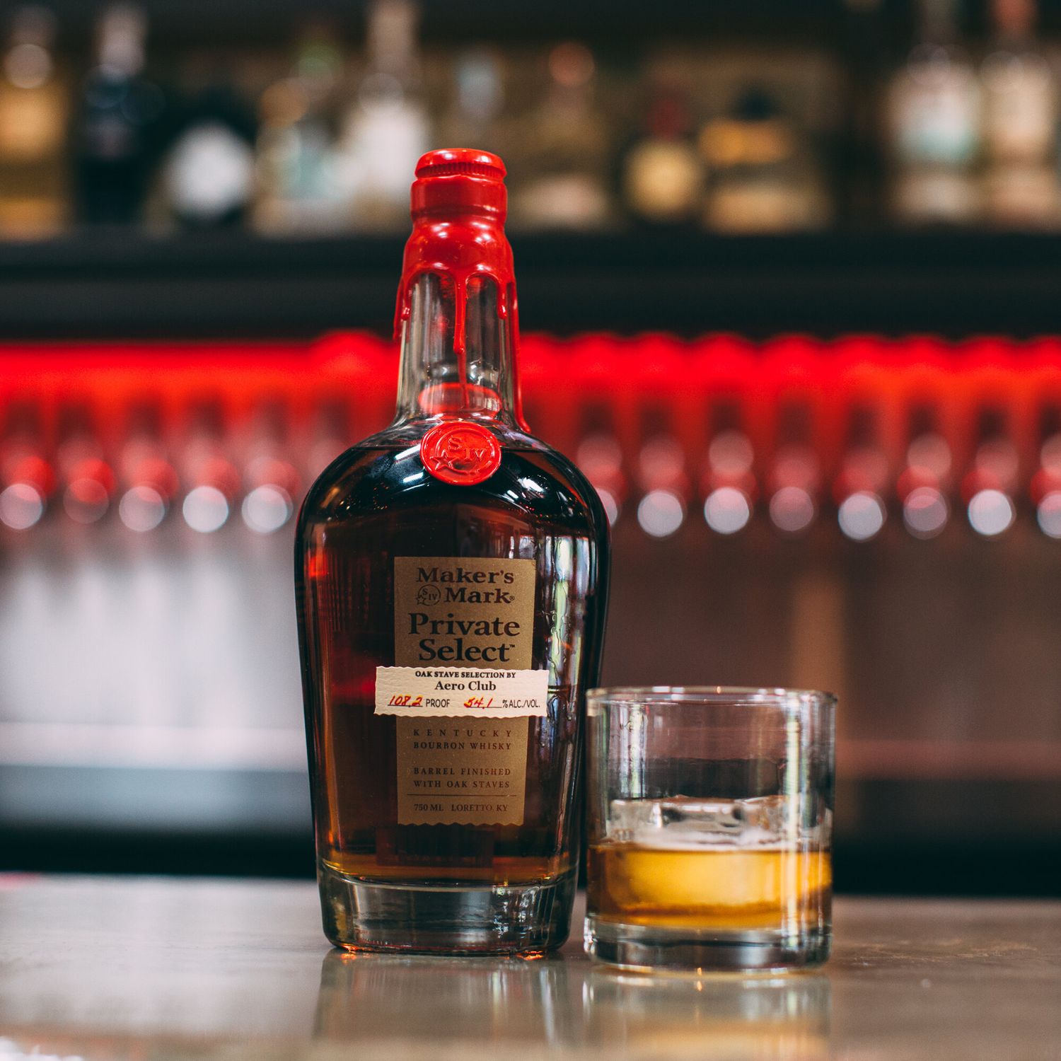 Maker's Mark Private Select bourbon bottle beside a glass of whiskey on a bar counter.