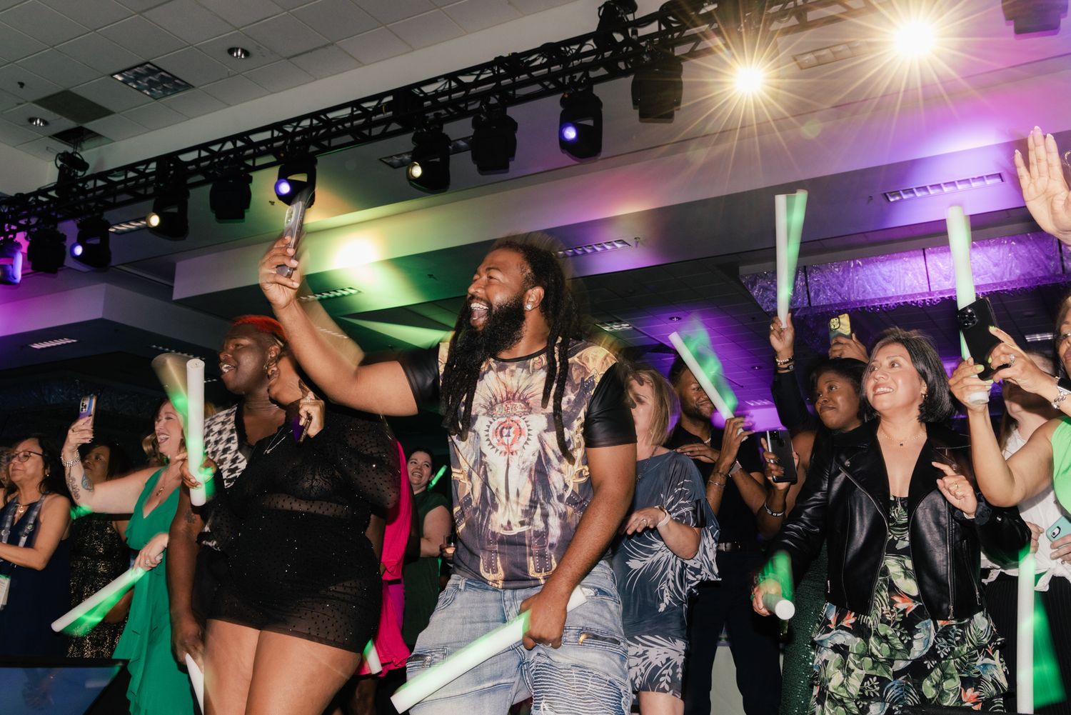 Excited crowd dancing and holding glow sticks at a vibrant indoor event with colorful stage lighting.