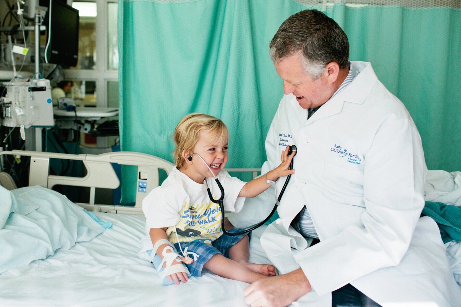 A smiling child uses a stethoscope on a doctor in a white coat during a hospital visit.