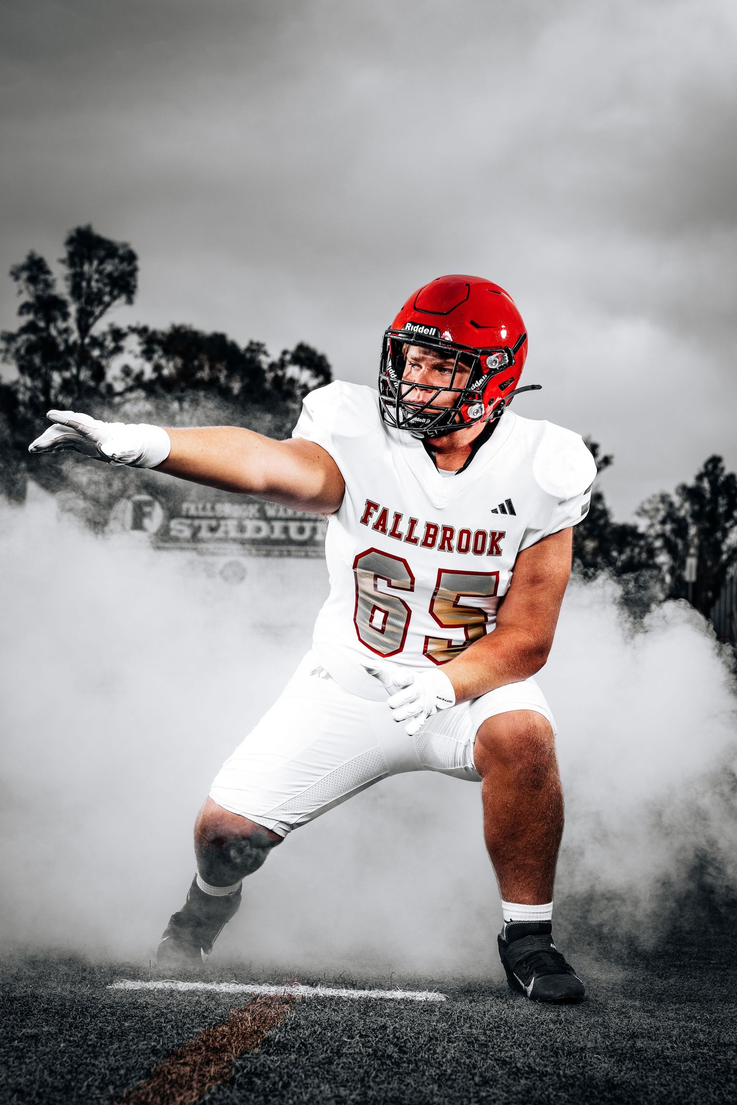 Football player in red helmet and white Fallbrook jersey emerges dramatically through smoke on the field.
