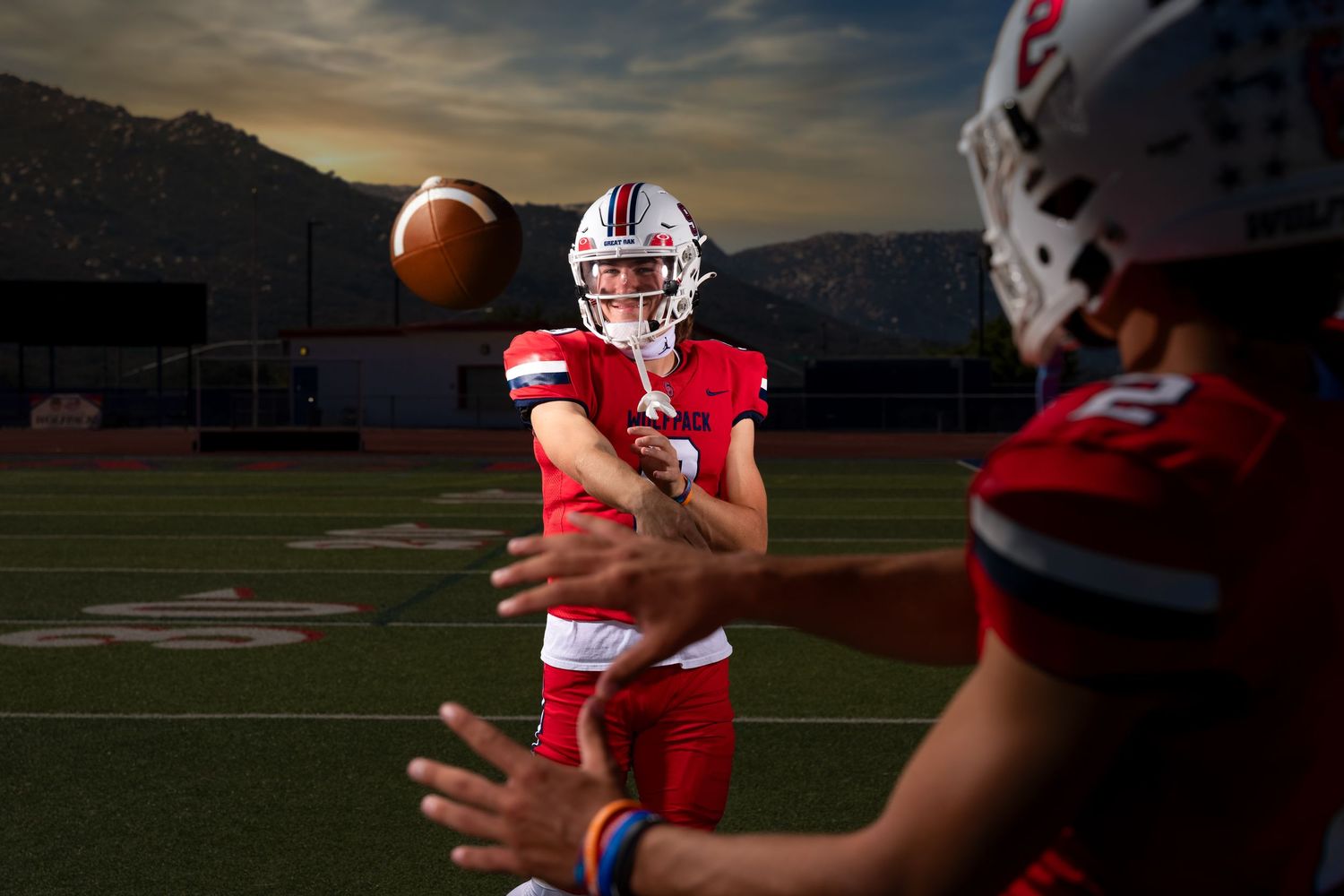 Football players in red and white uniforms practice on field during dramatic sunset with dark clouds overhead.