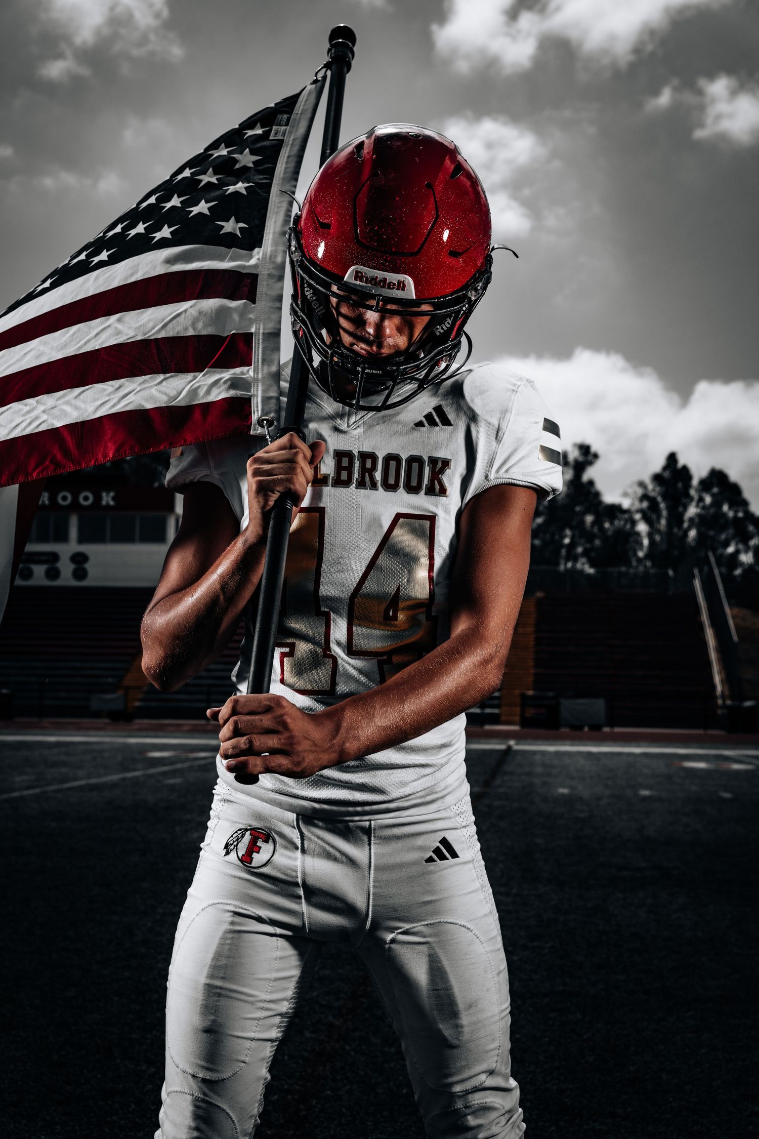 American football player in white uniform holds flag against dramatic black and white sky with selective red coloring.
