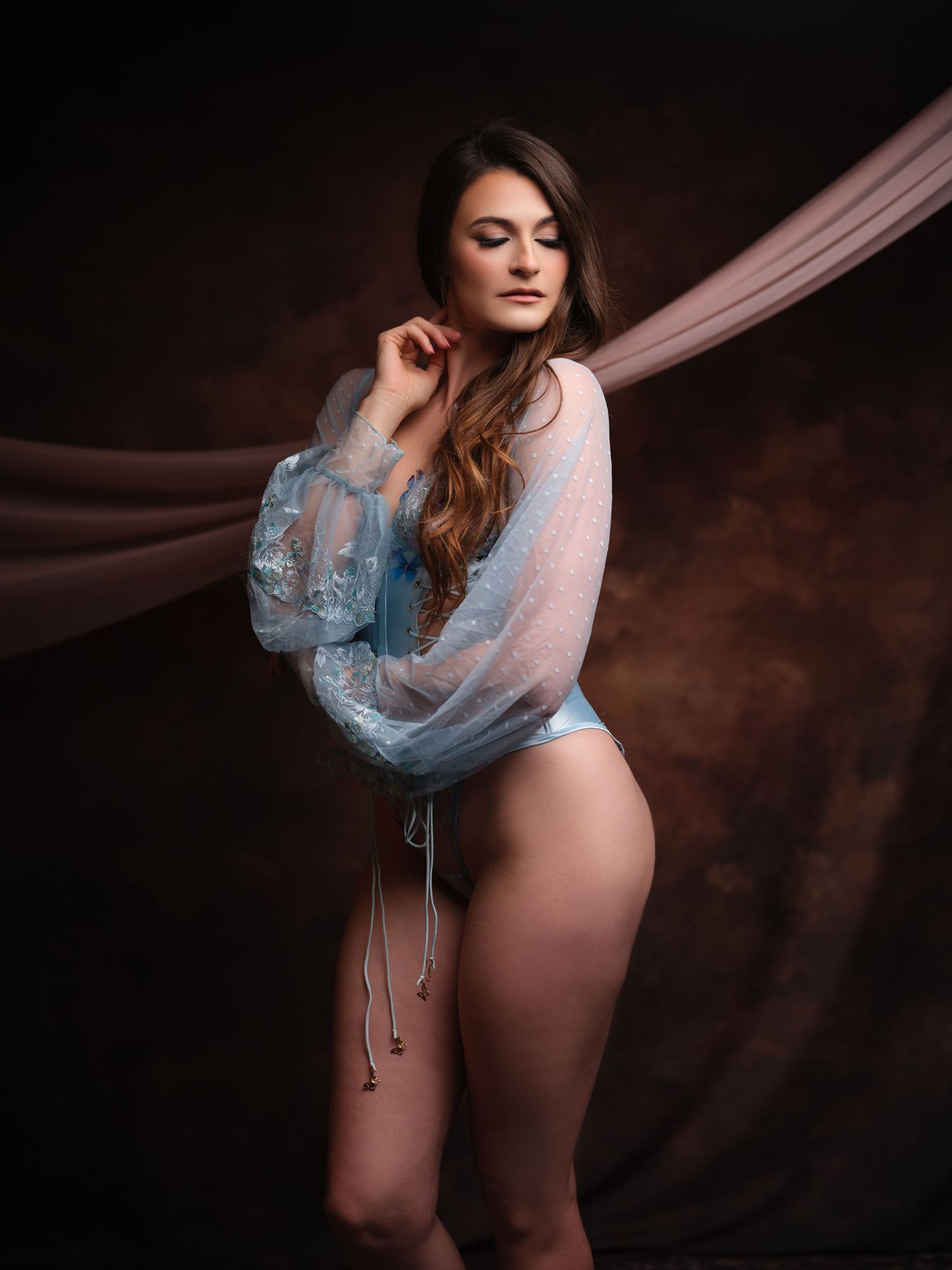 Woman in sheer blue top and matching bottoms poses gracefully against a dark studio backdrop with draped fabric.