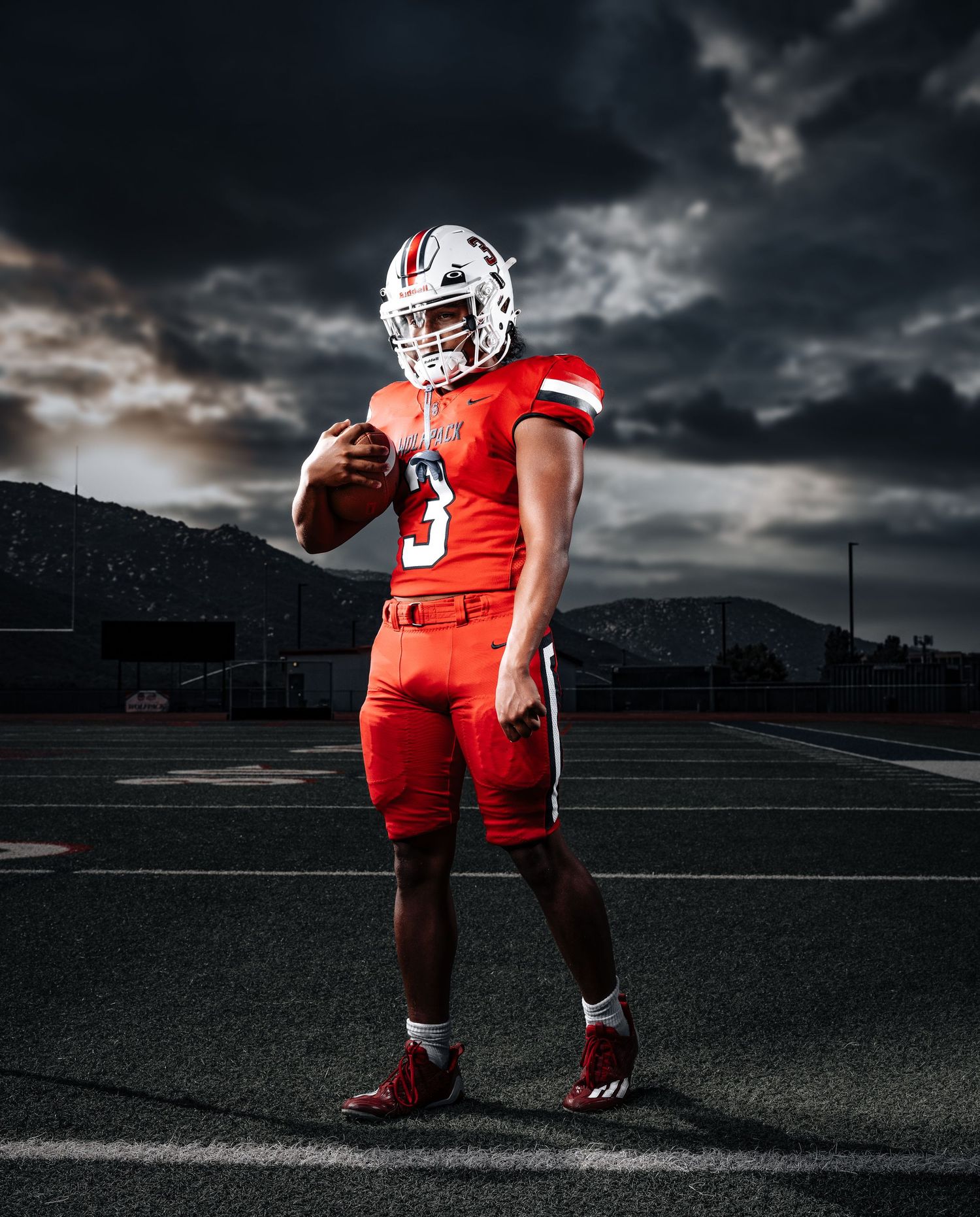 Football player in red uniform stands on dark field under dramatic stormy sky with selective lighting effects.