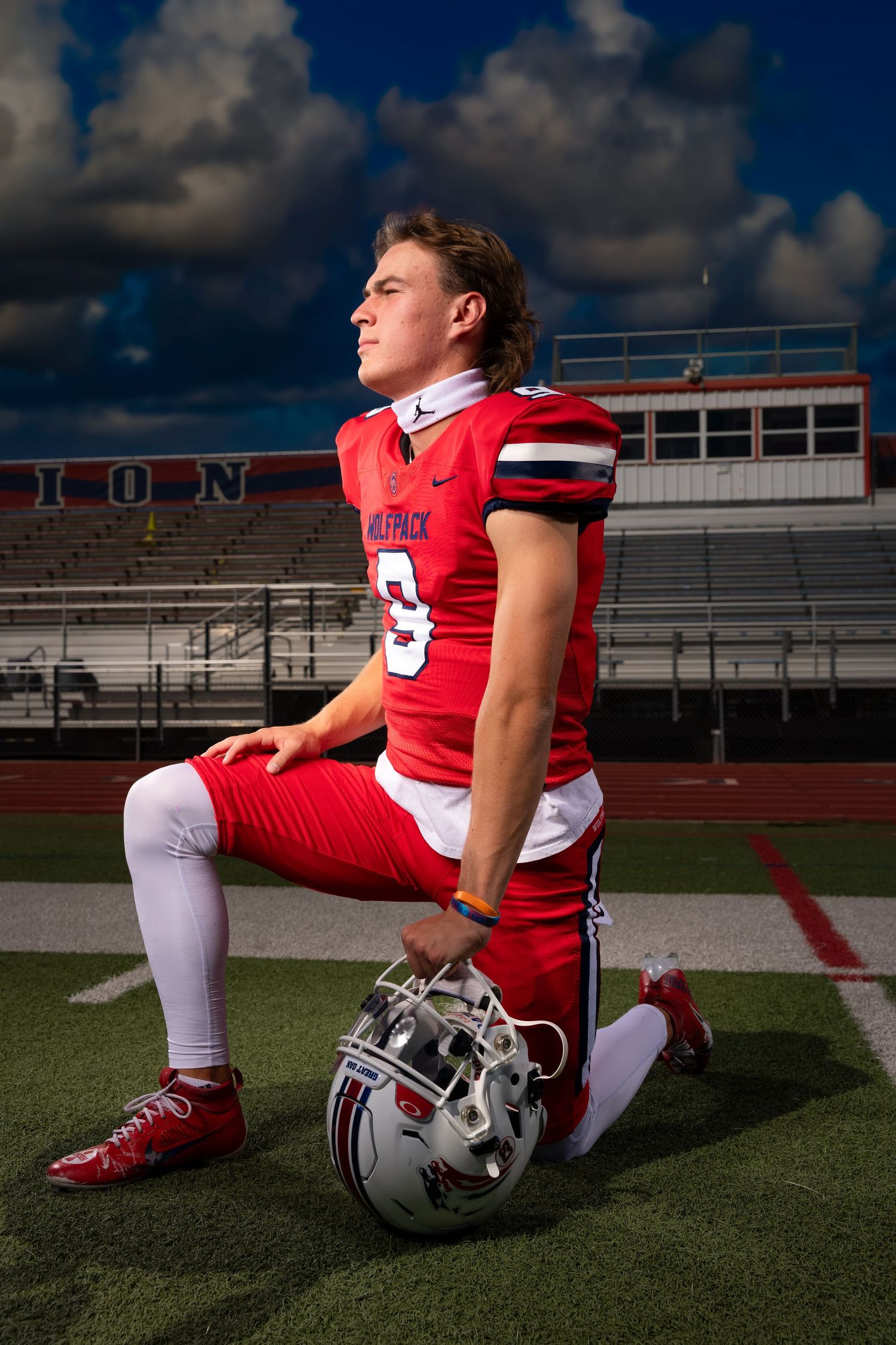 Football player in red and white uniform sits on field with helmet beside them during dramatic outdoor photoshoot.