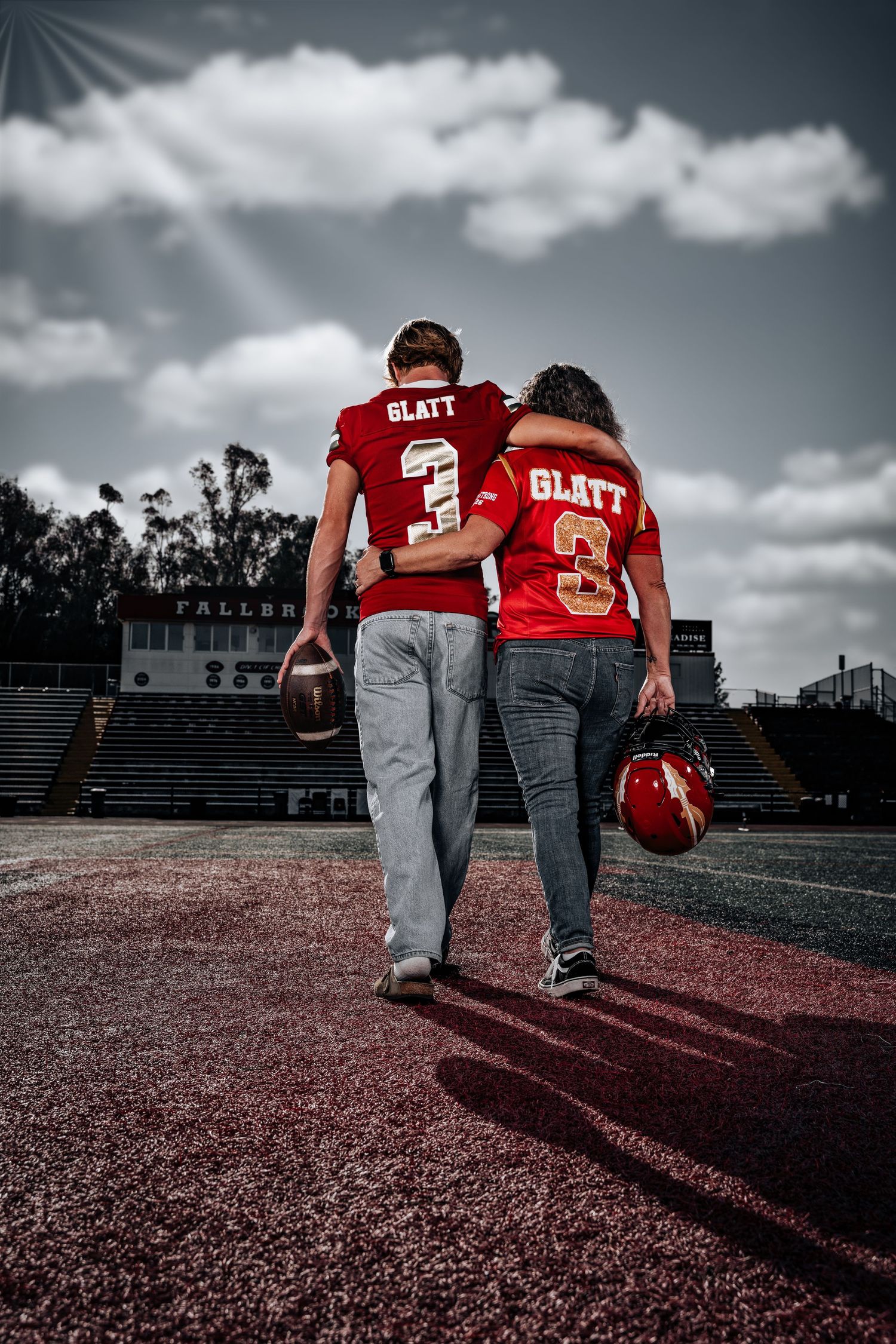 Two football players in red jerseys walk away across a field under dramatic cloudy skies in a cinematic composition.