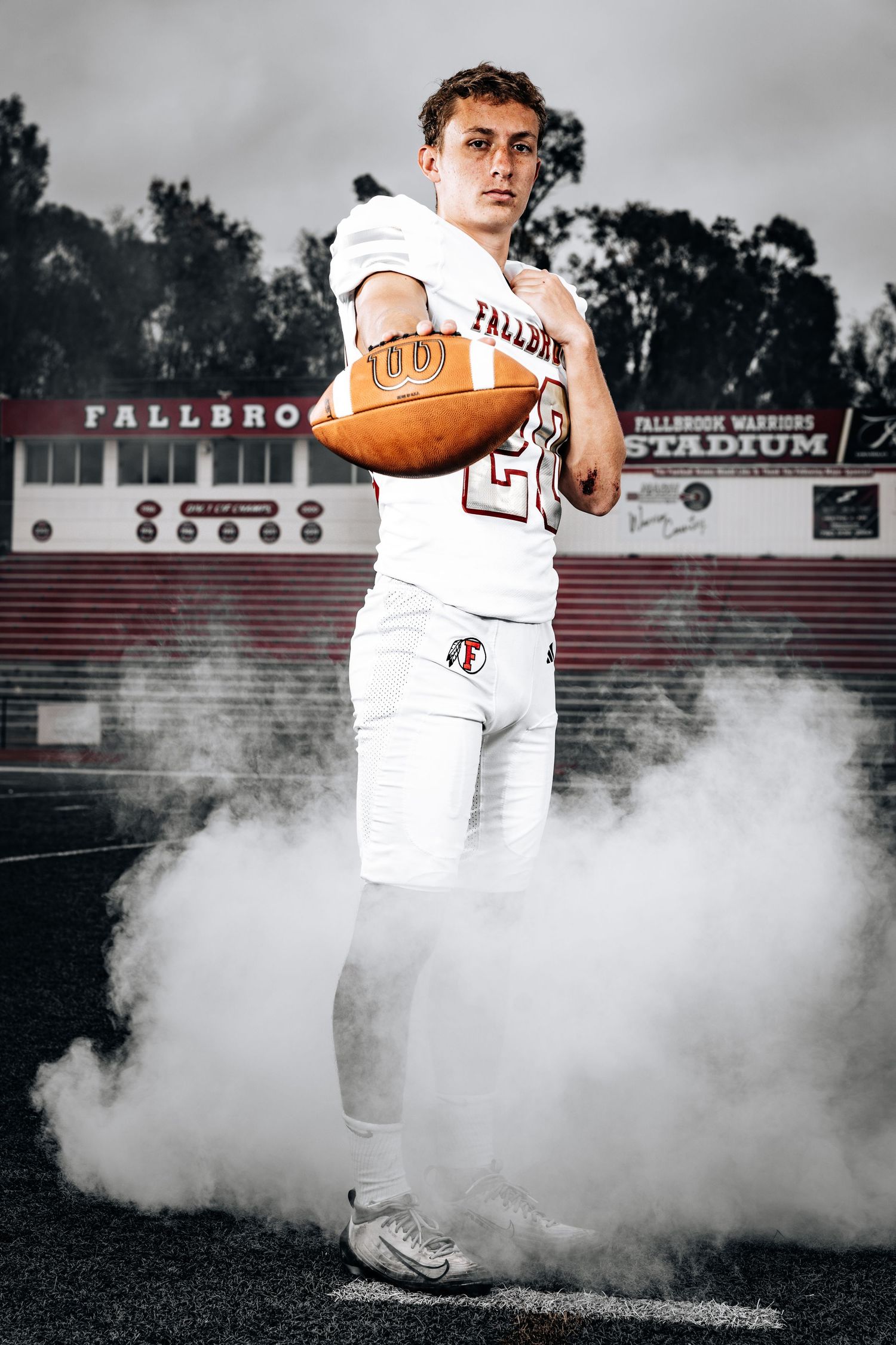 Football player in white uniform emerges dramatically through smoke on field during atmospheric sports photoshoot.