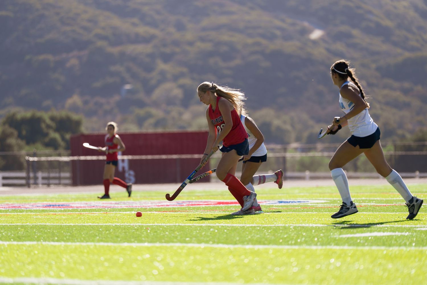 Athletes stretch on a sunny sports field with mountains visible in the background.