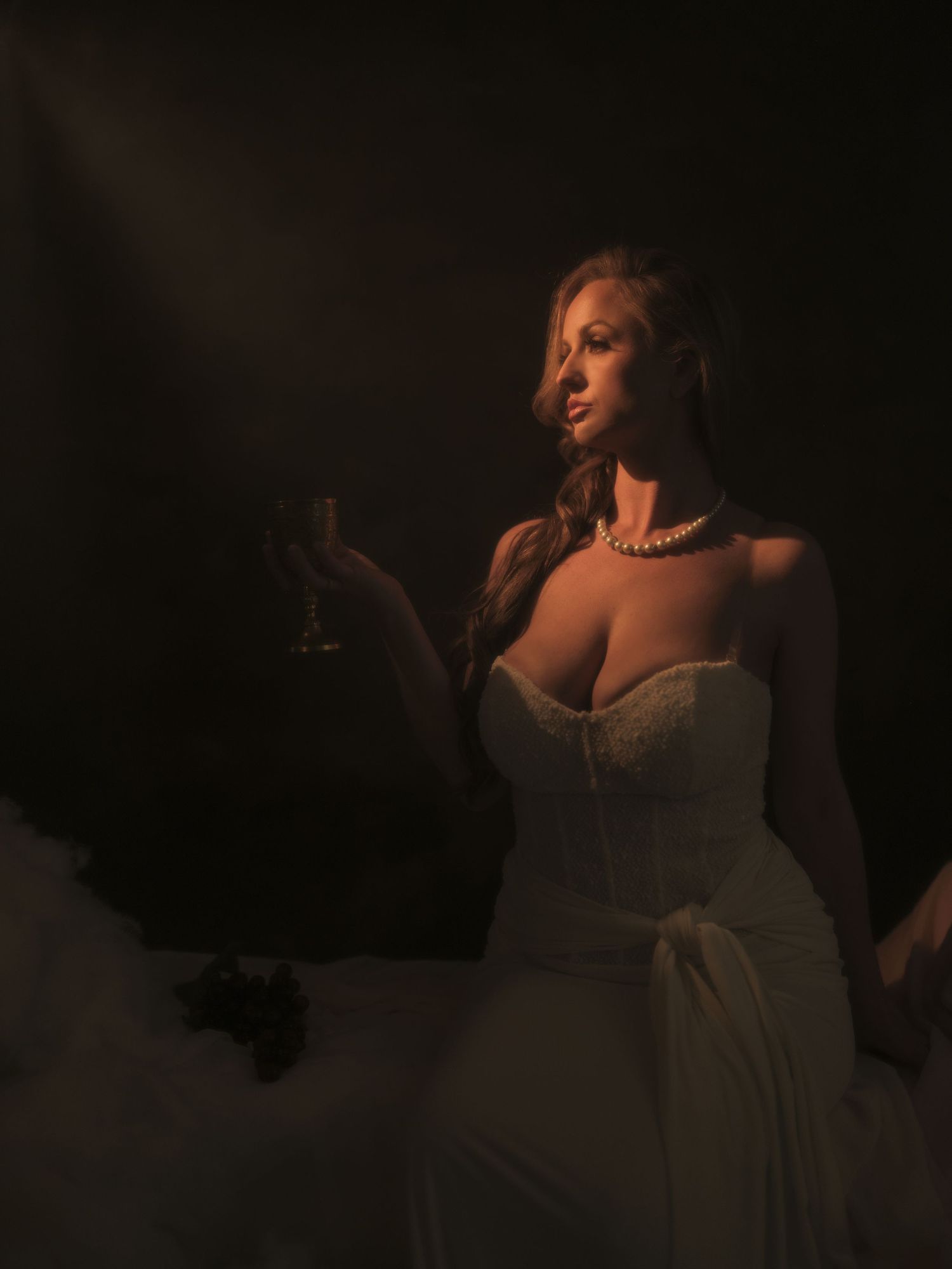 Elegant blonde woman in white corset gown holding wine glass, pearl necklace, dramatic moody lighting.