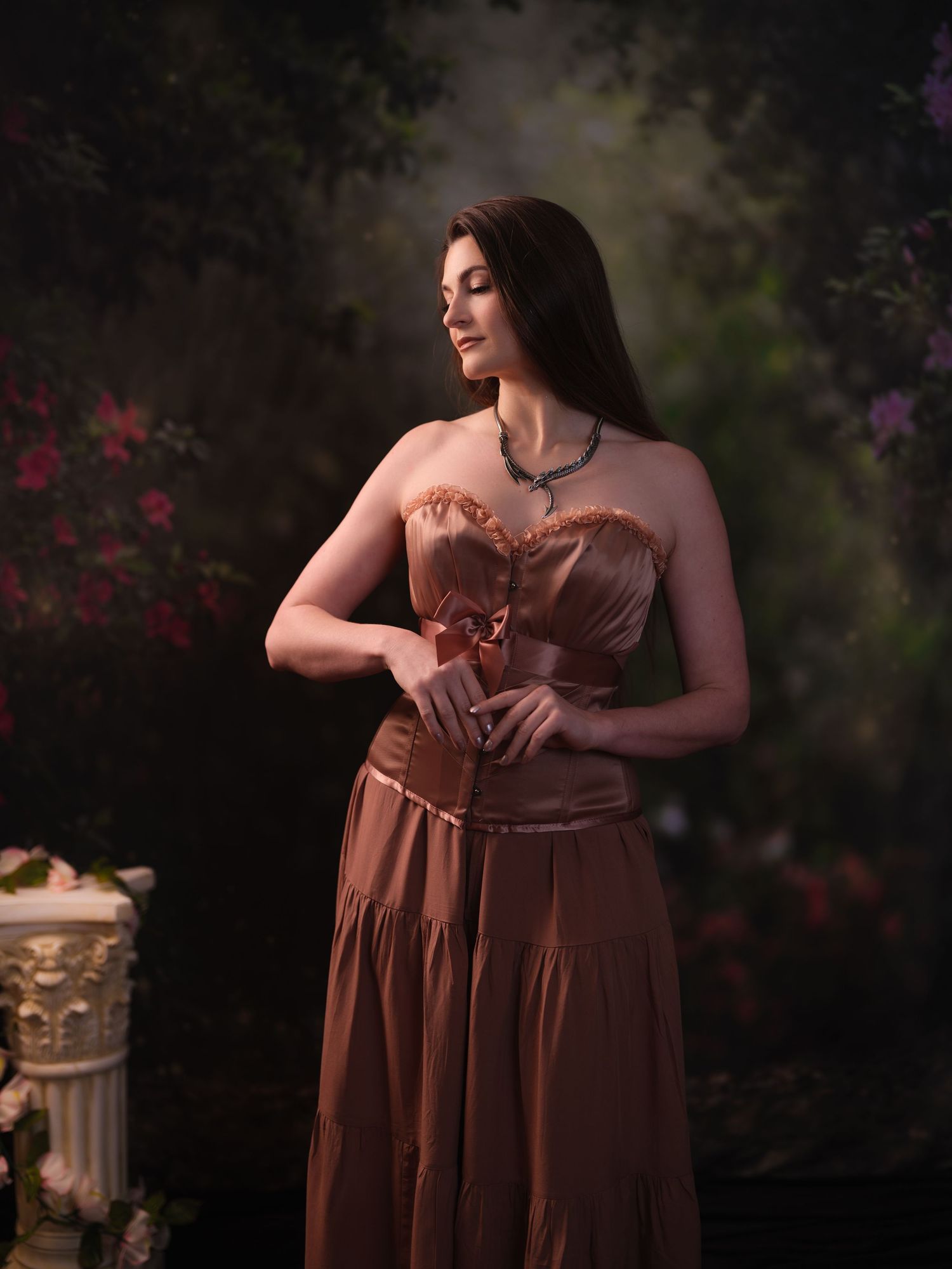 Romantic outdoor portrait in brown flowing dress against dark garden setting with flowers.