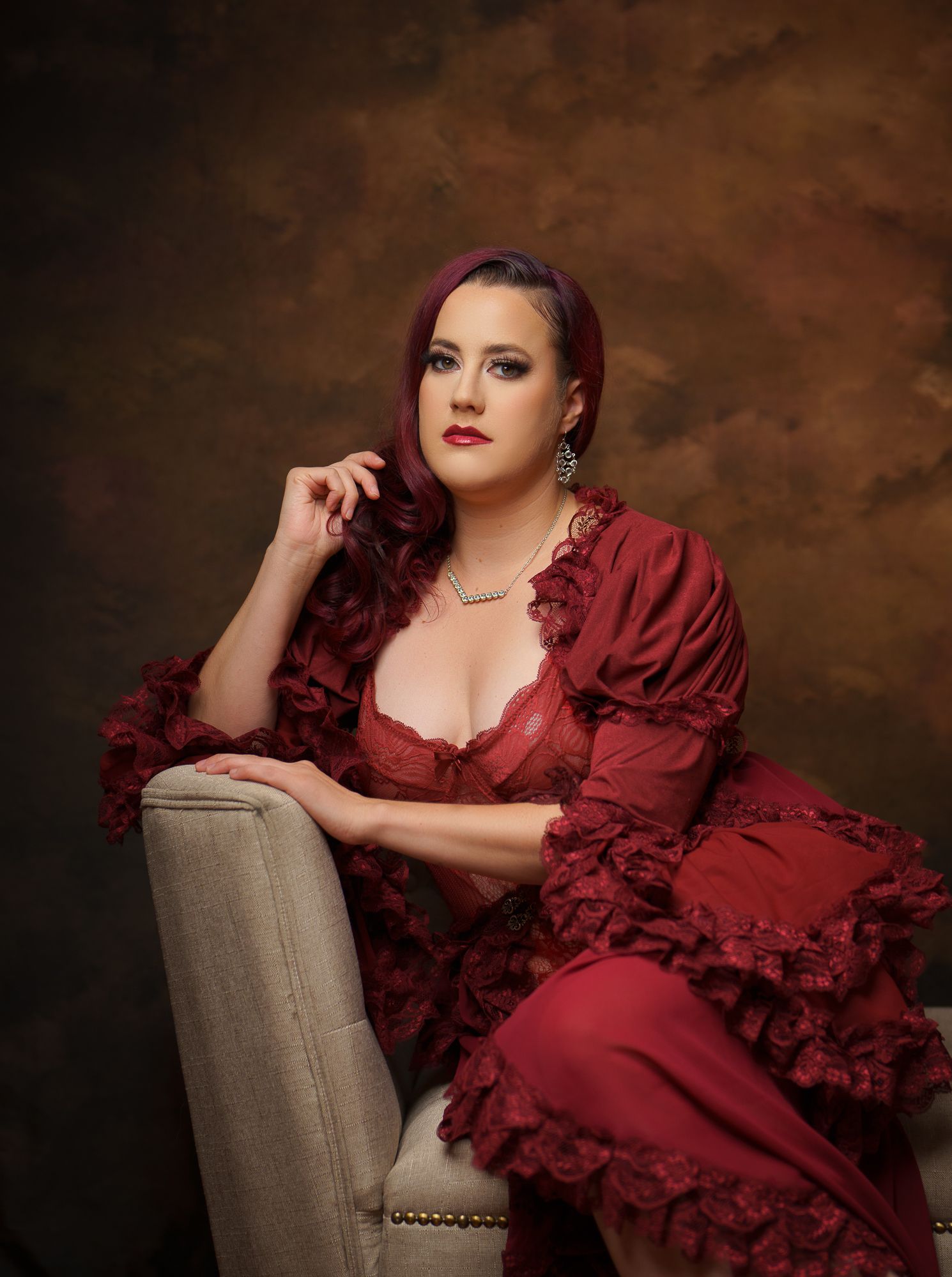 Glamour portrait series in flowing red dress posed on grey upholstered chair.