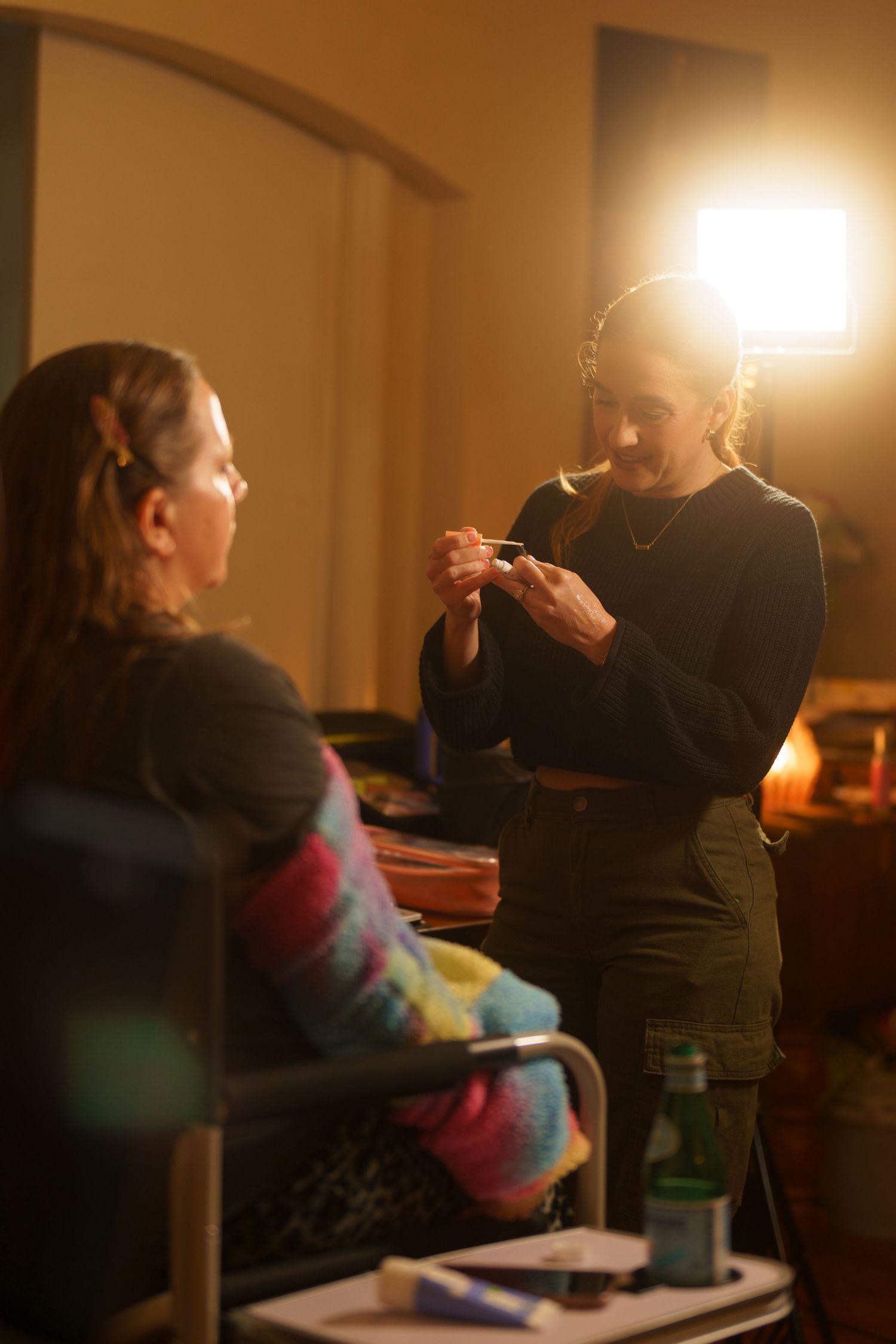 A makeup artist applies cosmetics to a seated woman in a warmly lit room during a behind-the-scenes session.