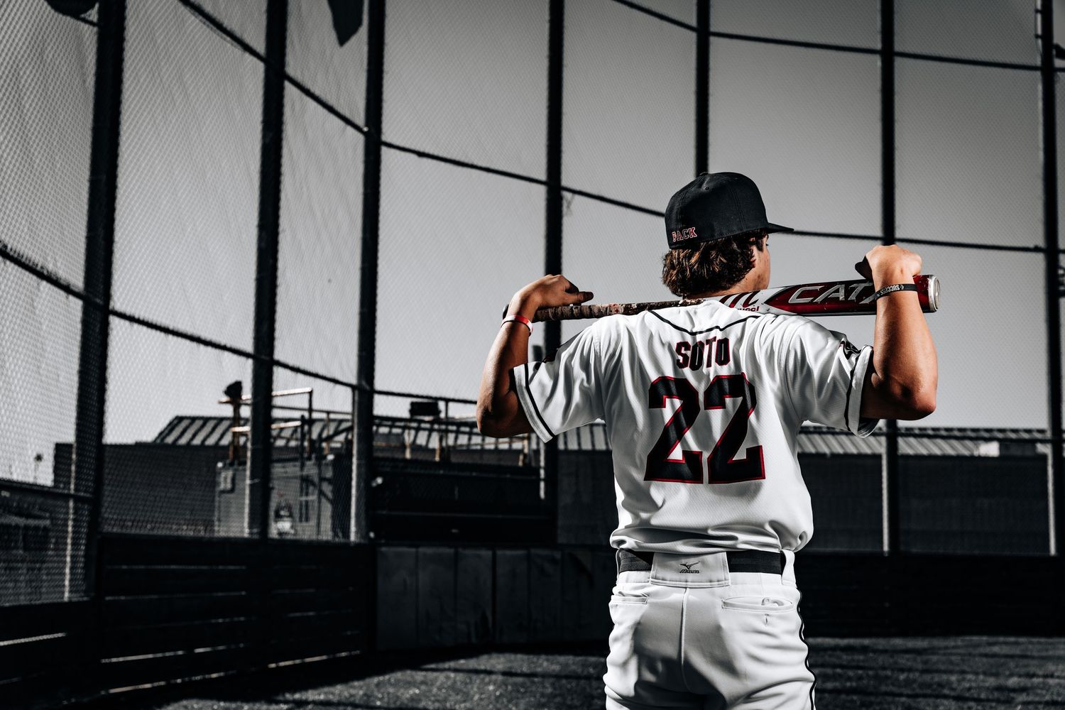 Baseball player in white uniform swings bat during game with stadium backdrop and wire netting visible.